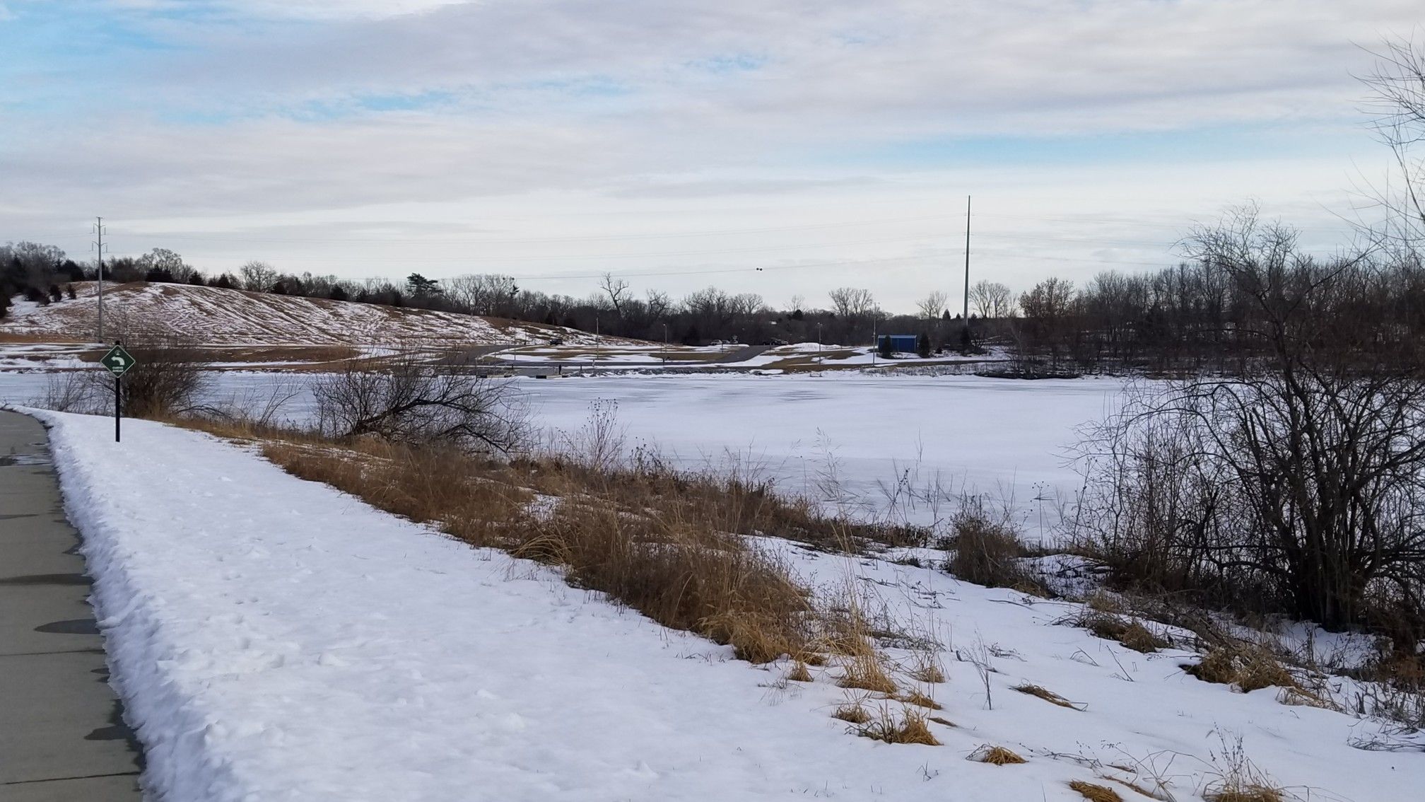 Snow-covered paved path next to a frozen pond with hills. - Cunningham Lake Bridge loop from Glenn Cunningham Lake Park