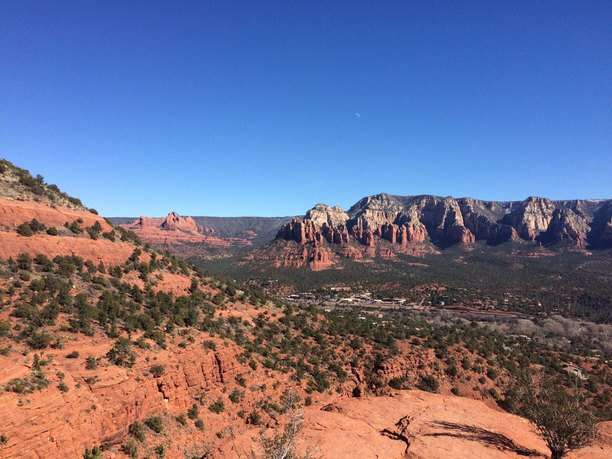Vast desert landscape with red rock formations, green valleys, and distant mountains under clear sky. - Brewer Trail