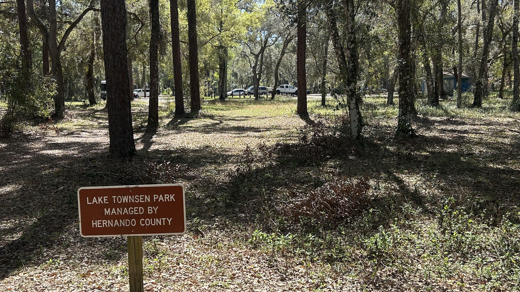 A brown wooden sign in a sun-dappled forest with parked vehicles. - Withlacoochee State Trail – Floral City, Florida loop from Floral City