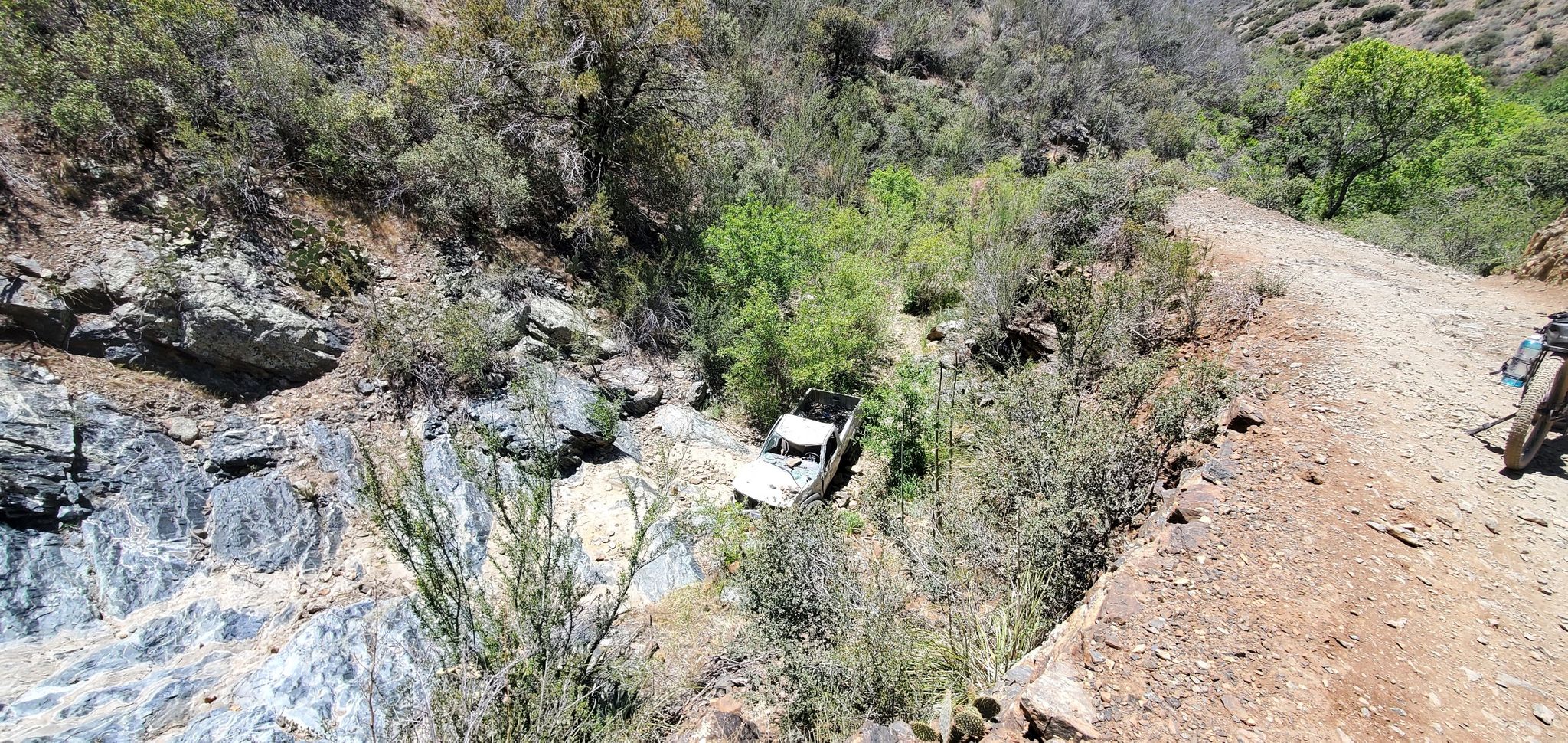 A white wrecked vehicle lies in a rocky, brushy ravine under bright sun. - Indian Mesa Trail