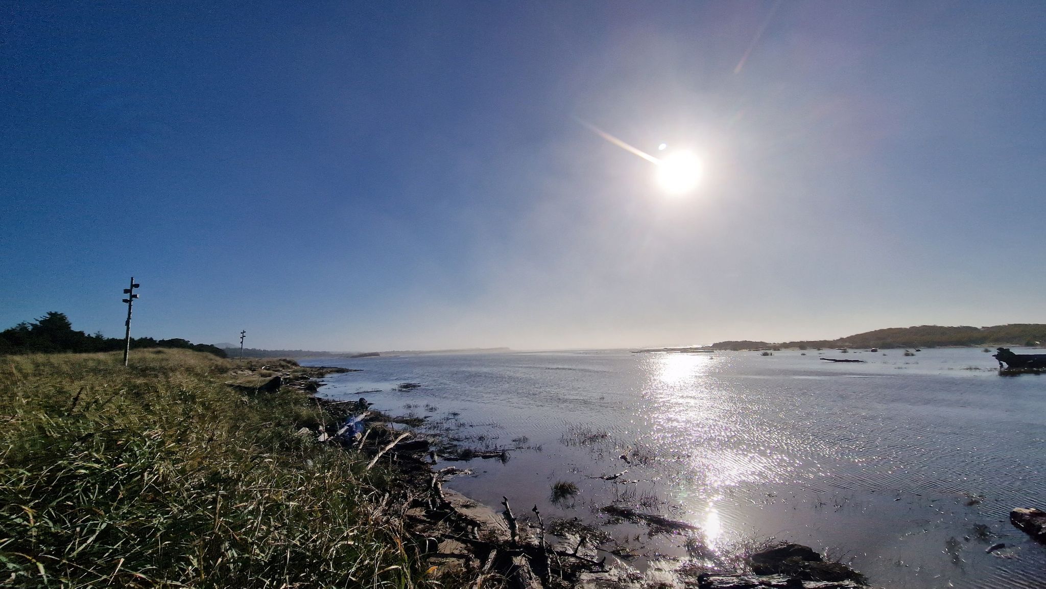 Bright sun over a calm bay with grassy shore and distant boats. - Siltcoos River Paddle Route