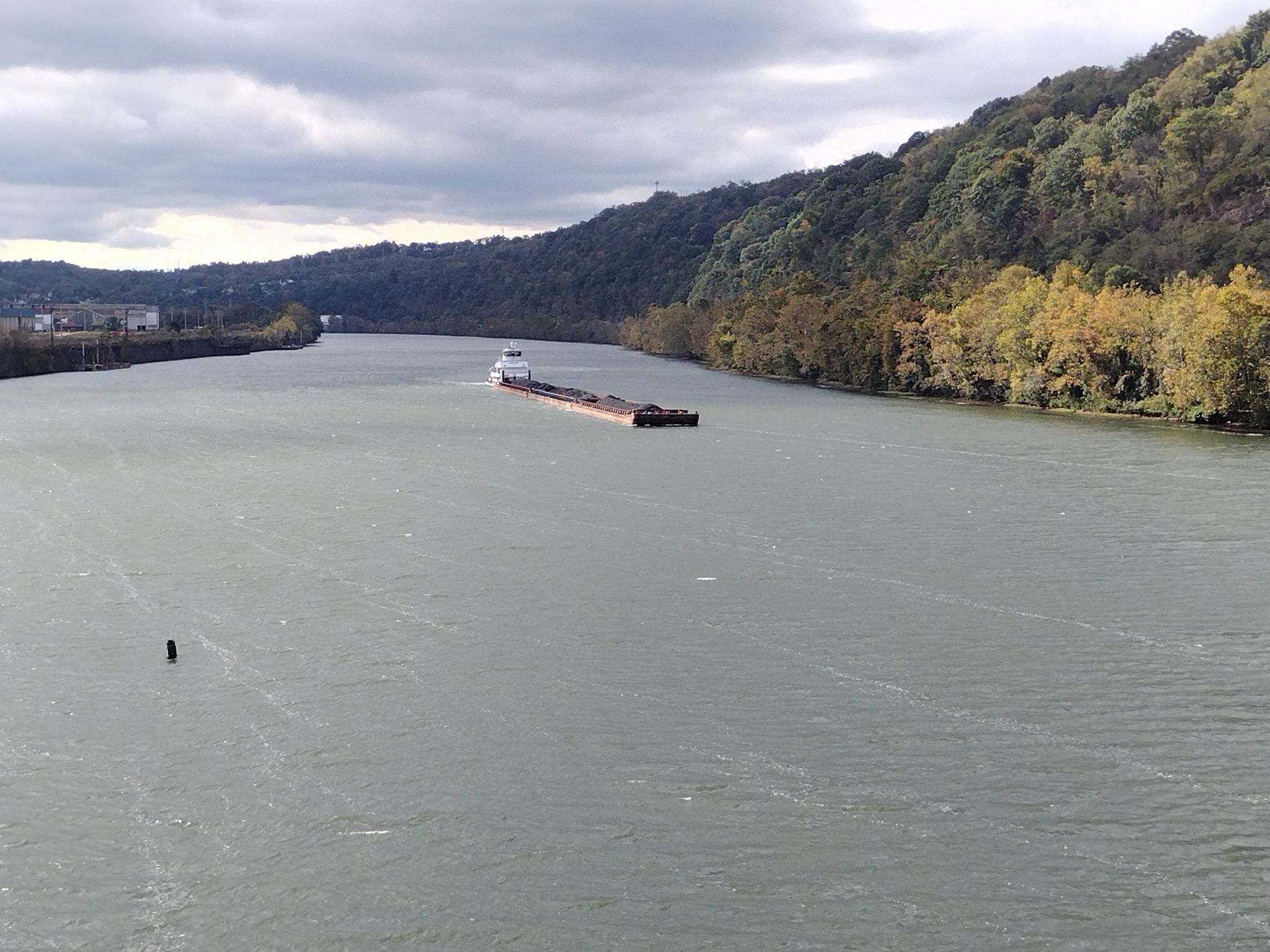 A long barge floats on a wide river, flanked by forested hills, cloudy sky. - McDonald Trestle Bridge – National Tunnel loop from Steel Plaza