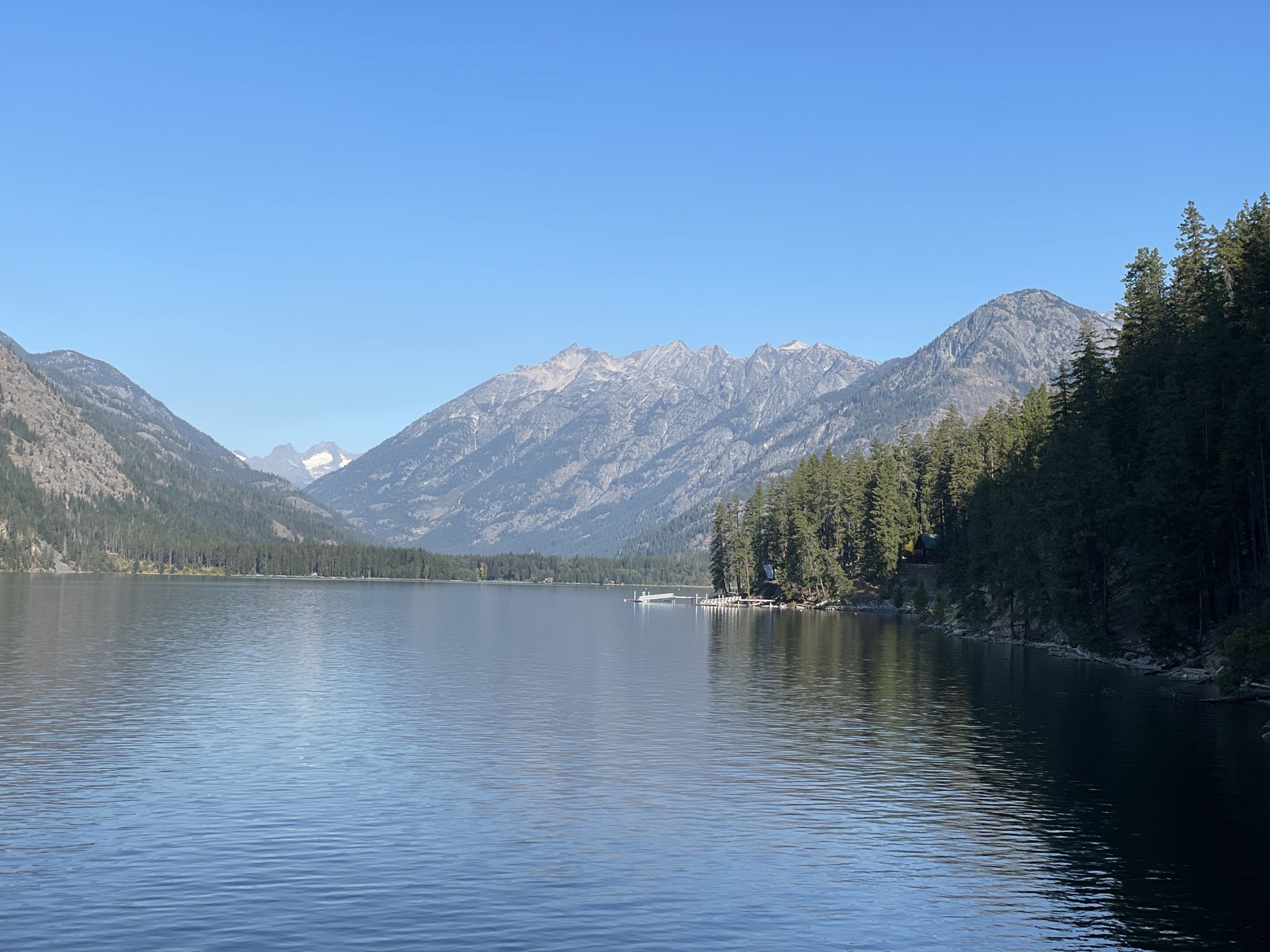 A serene lake surrounded by forested mountains under a clear blue sky. - Chelan Lakeshore Trail