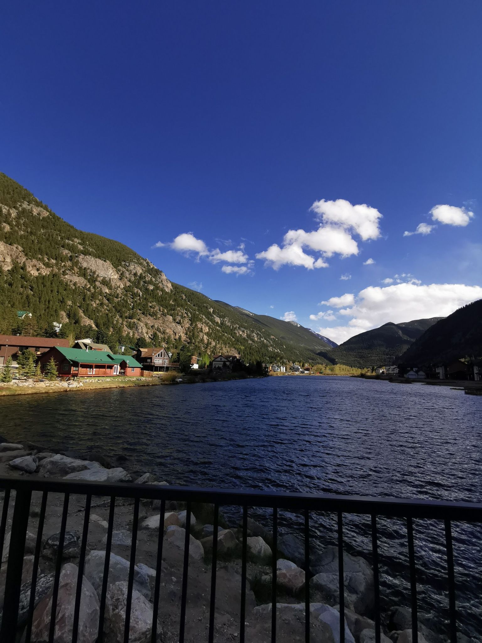 Lake with buildings along the shore, mountains, and a fence in foreground. - Tom Bennhoff Lake Trail loop