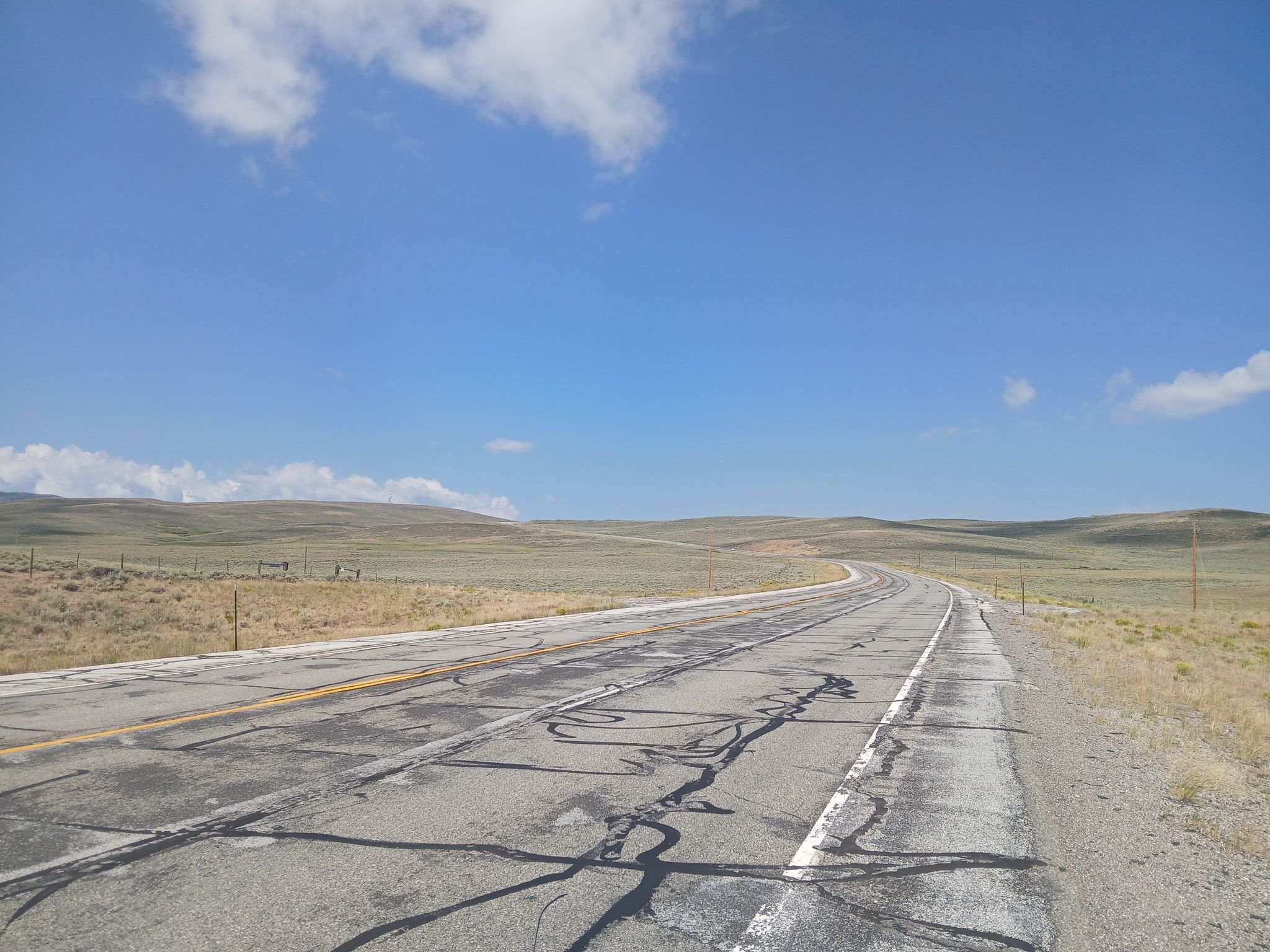 Cracked, winding road through dry, rolling hills under a bright blue sky. - TransAmerica Trail: Encampment to Colorado State Line