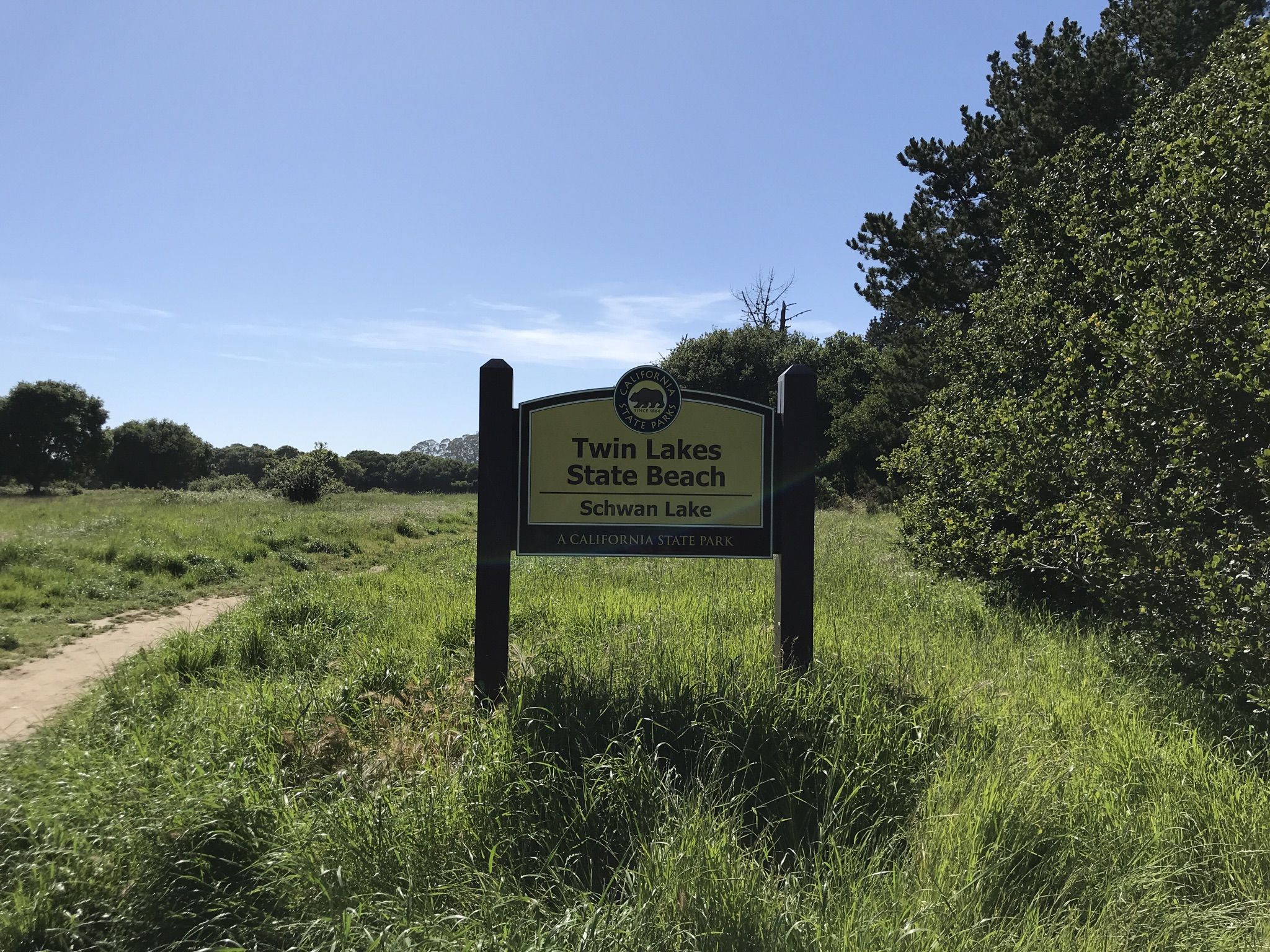 A wooden sign stands in a sunny, grassy field with a dirt path. - Schwan Lake Loop Trail
