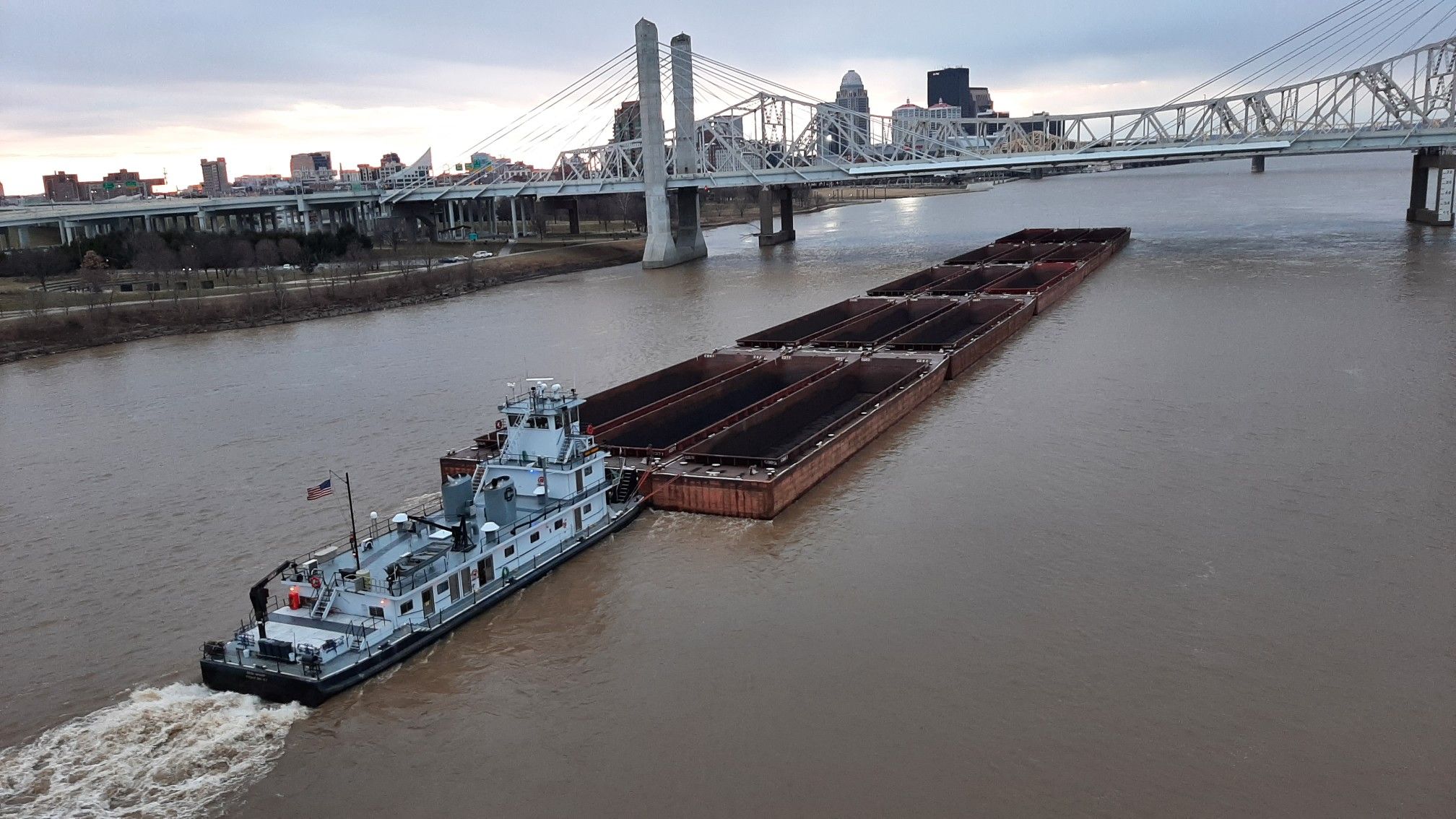A tugboat pushes barges on a wide river under a distant bridge. - Big Four Bridge – Big Four Station Park loop from Green Spring