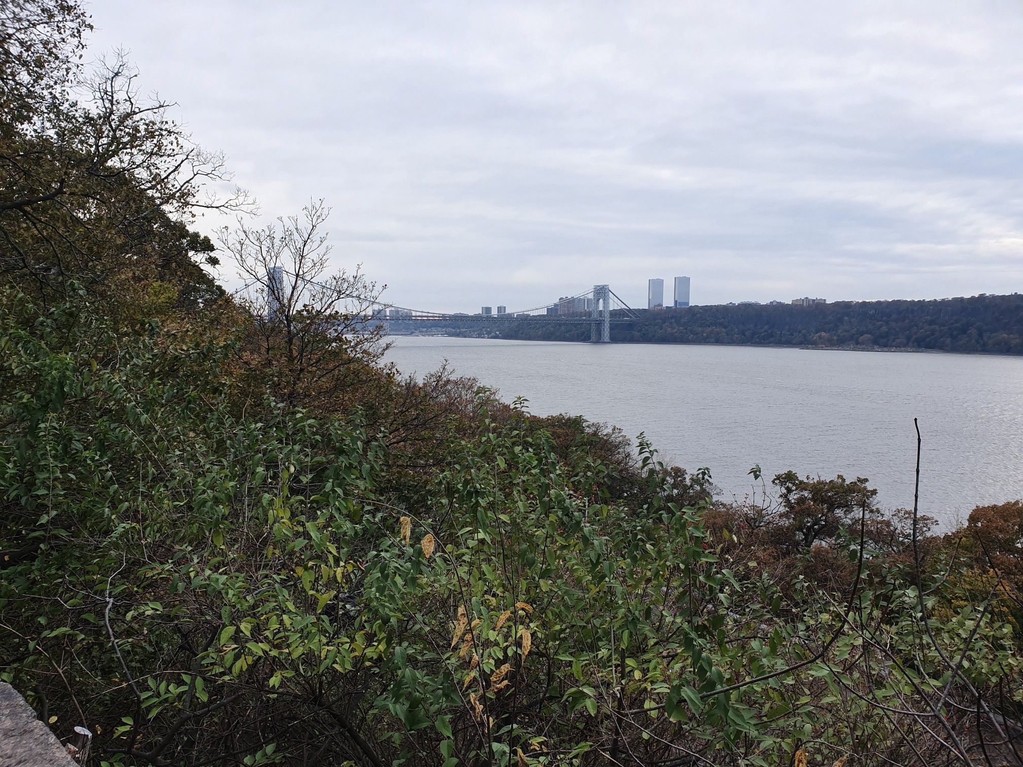 Overcast view of a wide river, distant bridge, and lush green foliage. - Fort Tryon Park Loop