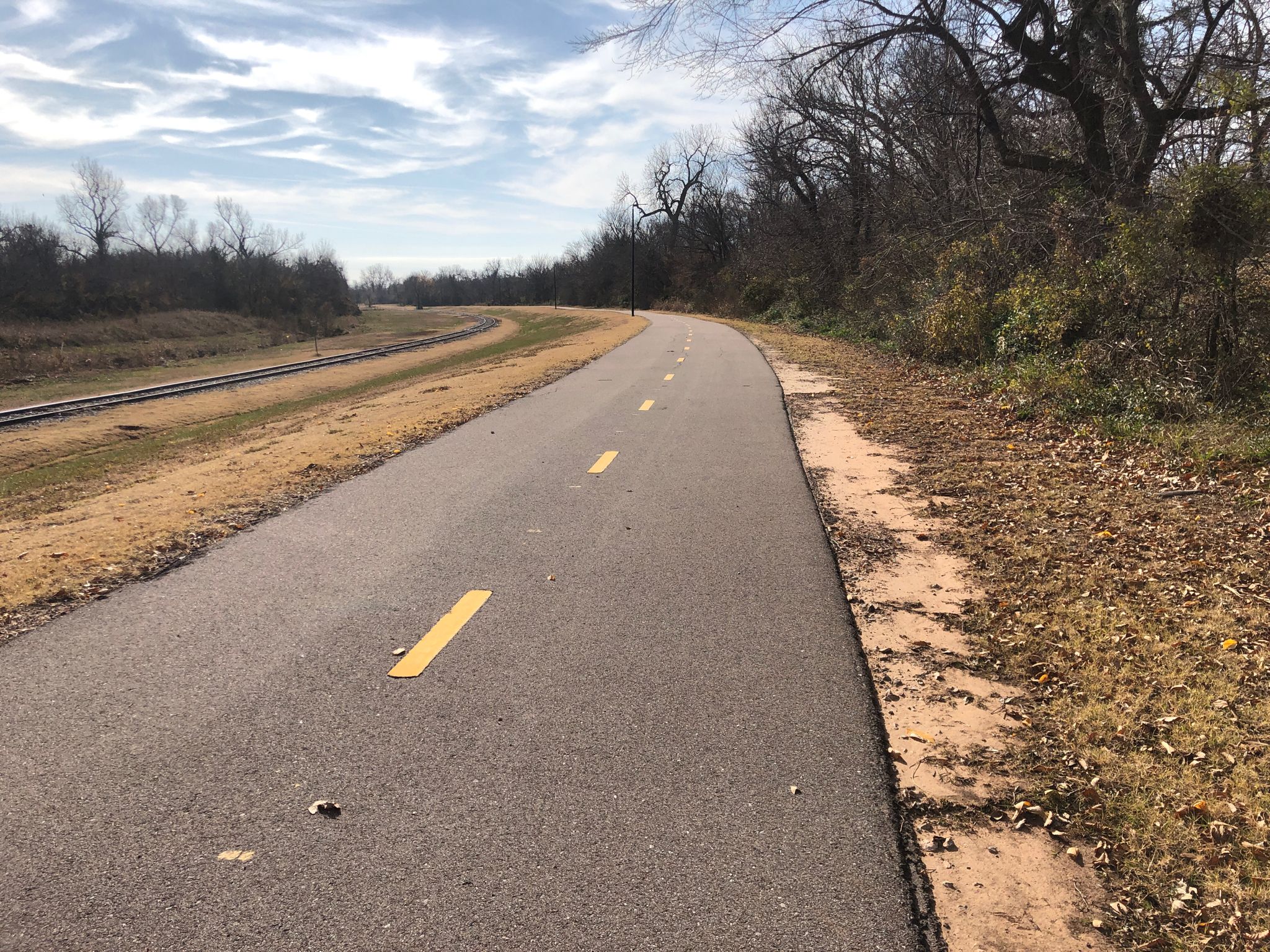 A paved path curves beside railroad tracks, with bare trees under a cloudy sky. - Oklahoma Railway Museum – Oklahoma Land Run Monument loop from Lake Aluma