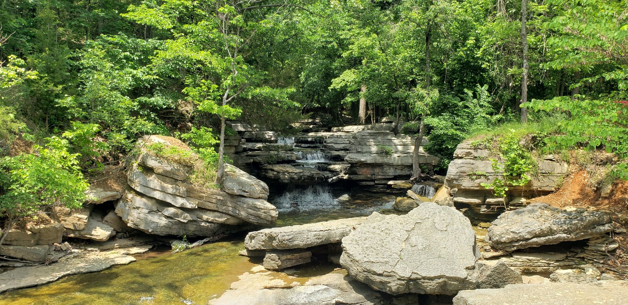 Sunny view of a small waterfall cascading over layered rocks in a green forest. - Back 40 Loop