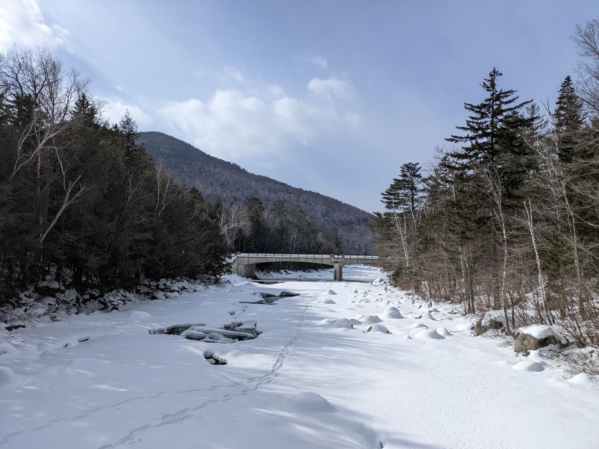 A snow-covered river flows under a bridge, surrounded by winter trees and mountains. - Lincoln Woods Trail