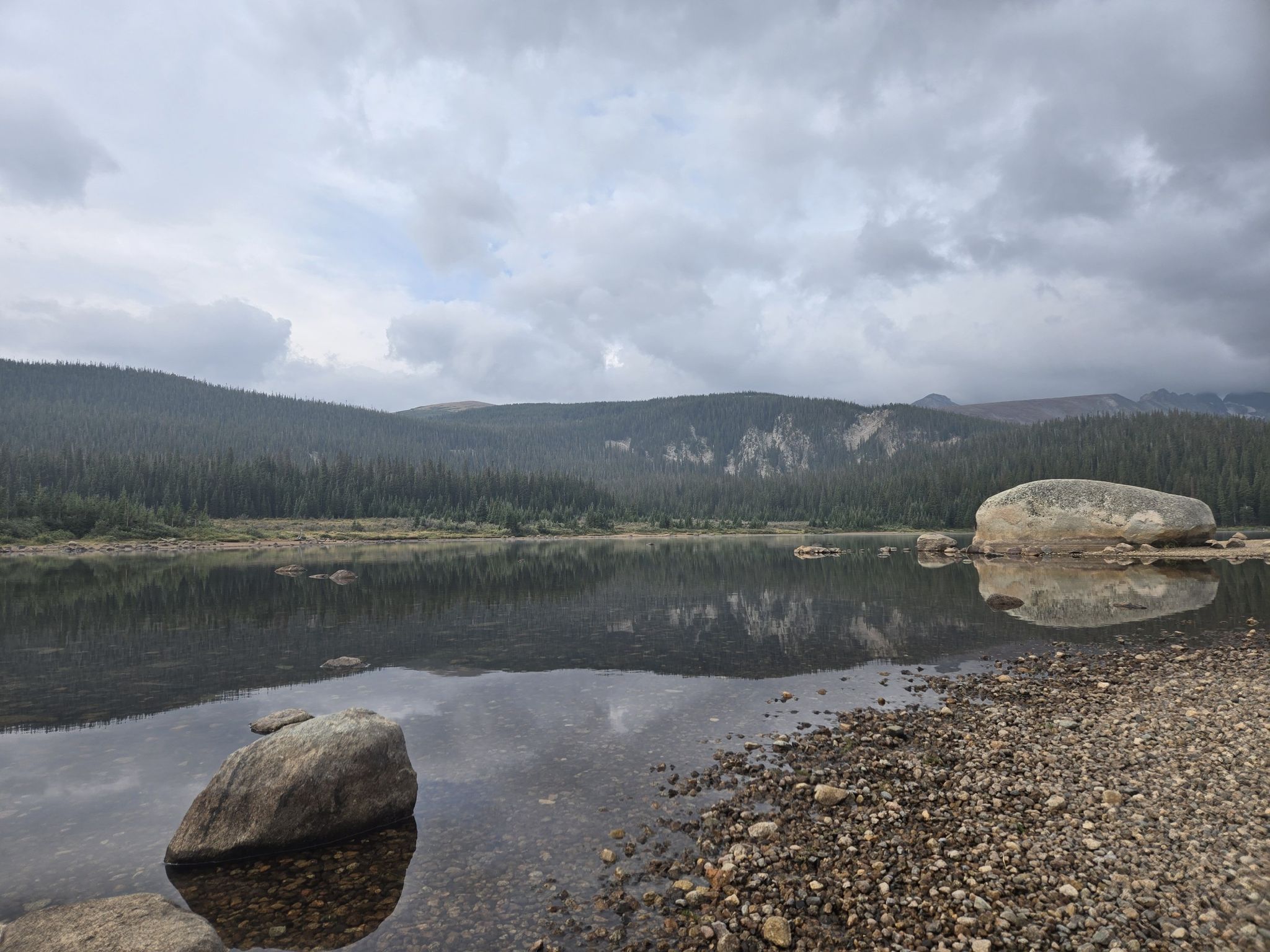 Overcast sky above a calm lake with forested hills and large rocks. - Ward and Brainard Lake via Lee Hill Drive from Pearl Street