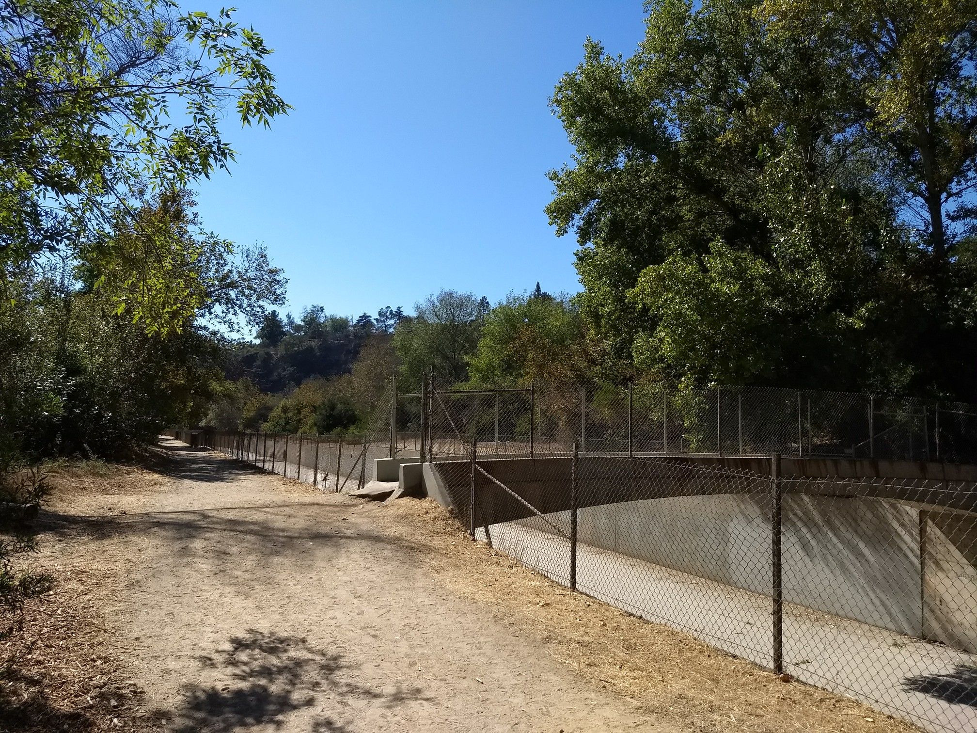 A dirt path runs beside a fenced concrete channel in a sunny, wooded landscape. - Lower Arroyo Seco Trail
