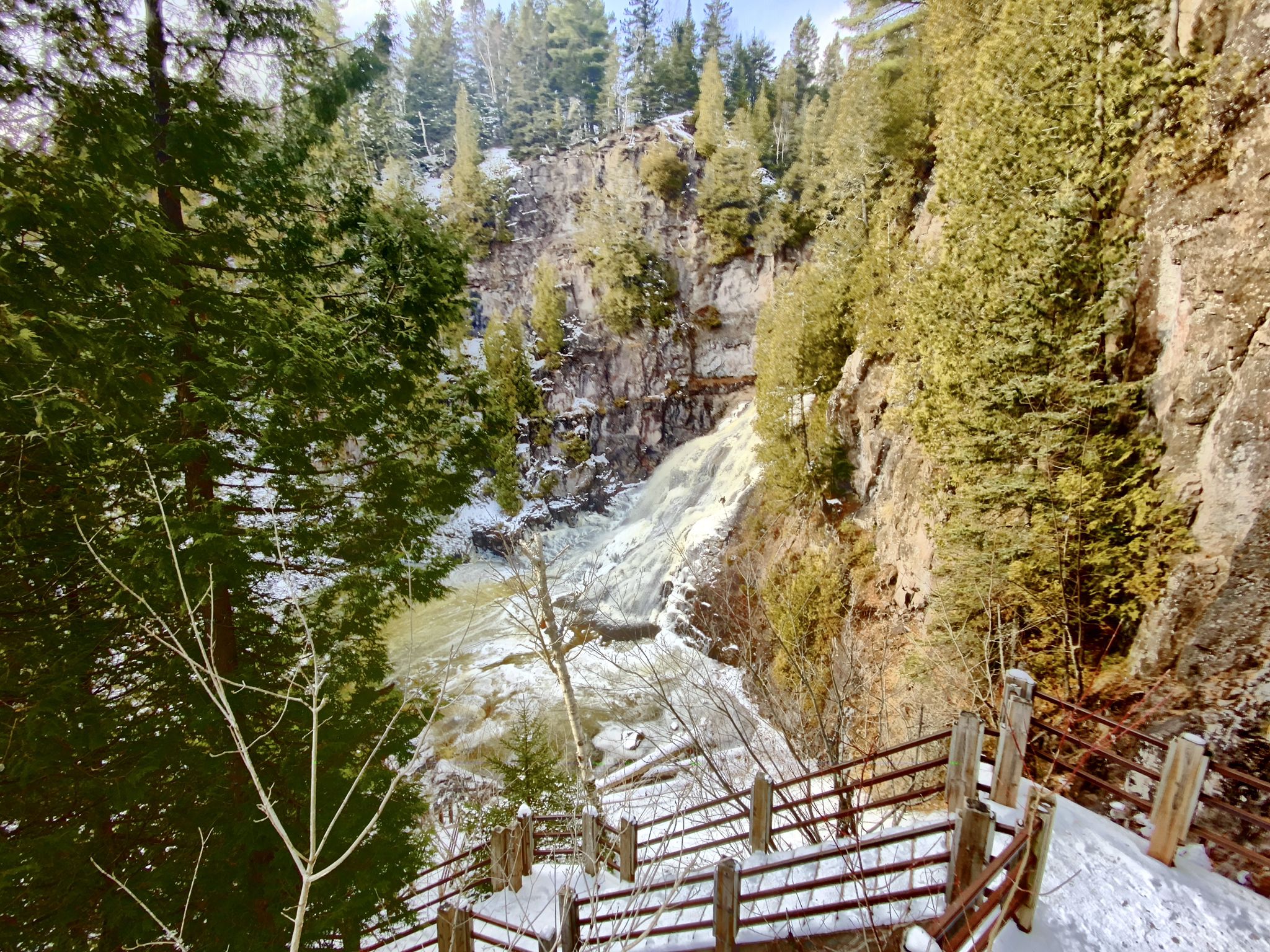 View of a waterfall in a snowy gorge, with wooden stairs and evergreens. - Caribou Falls via Superior Hiking Trail
