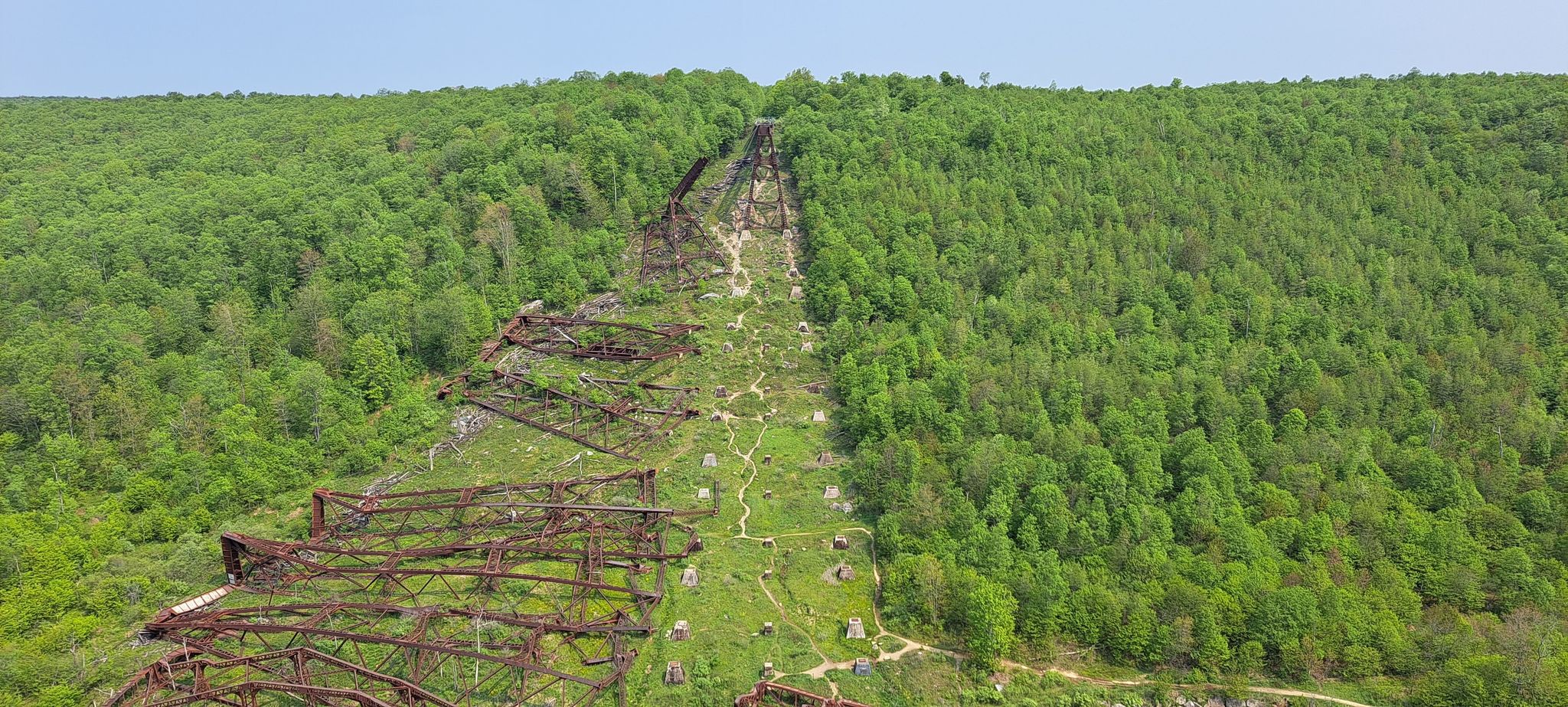 Collapsed metal bridge structure on a steep, lush green forested hillside under clear sky. - Kinzua Creek Trail