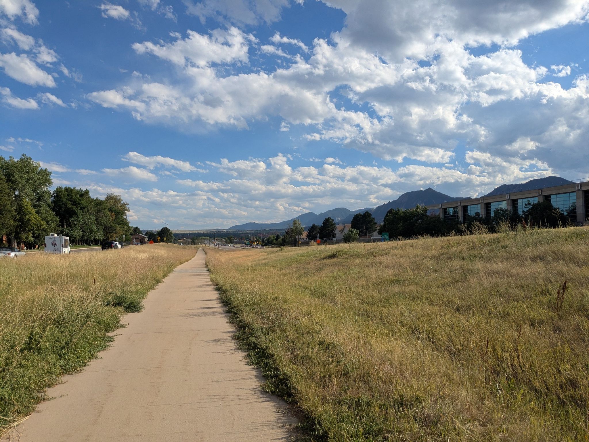 A paved path through dry grass fields, with mountains and buildings under a partly cloudy sky. - Ward General Store – Rapha Boulder Clubhouse loop from Cottonwood Trailhead