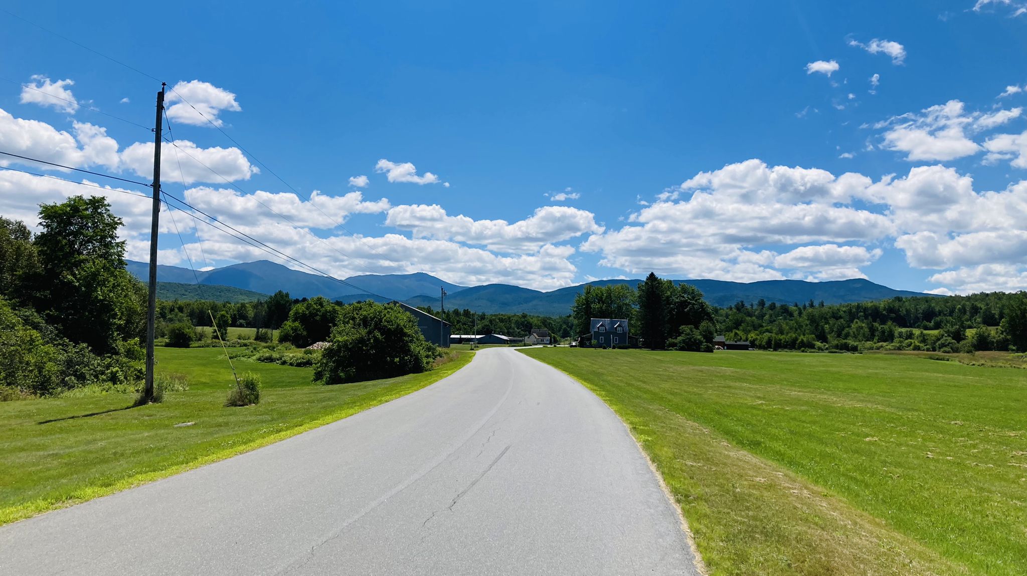 Paved road curving through green fields towards distant mountains under blue sky. - Cherry Mountain Loop