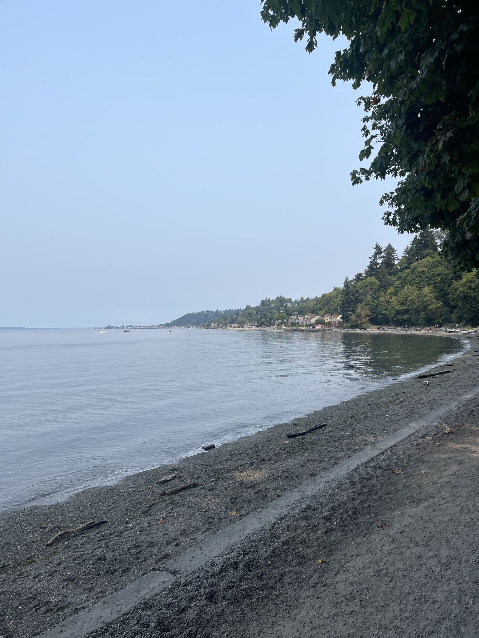 Hazy day at a calm pebble beach with distant forested shoreline. - Lowman Beach Park – Lincoln Park Trailhead loop from Lincoln Park