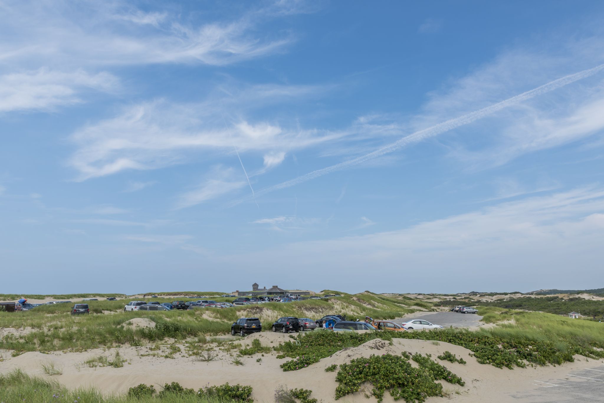 Expansive view of sandy, grassy dunes with distant buildings and cars under a blue sky. - Race Point Dunes Trail