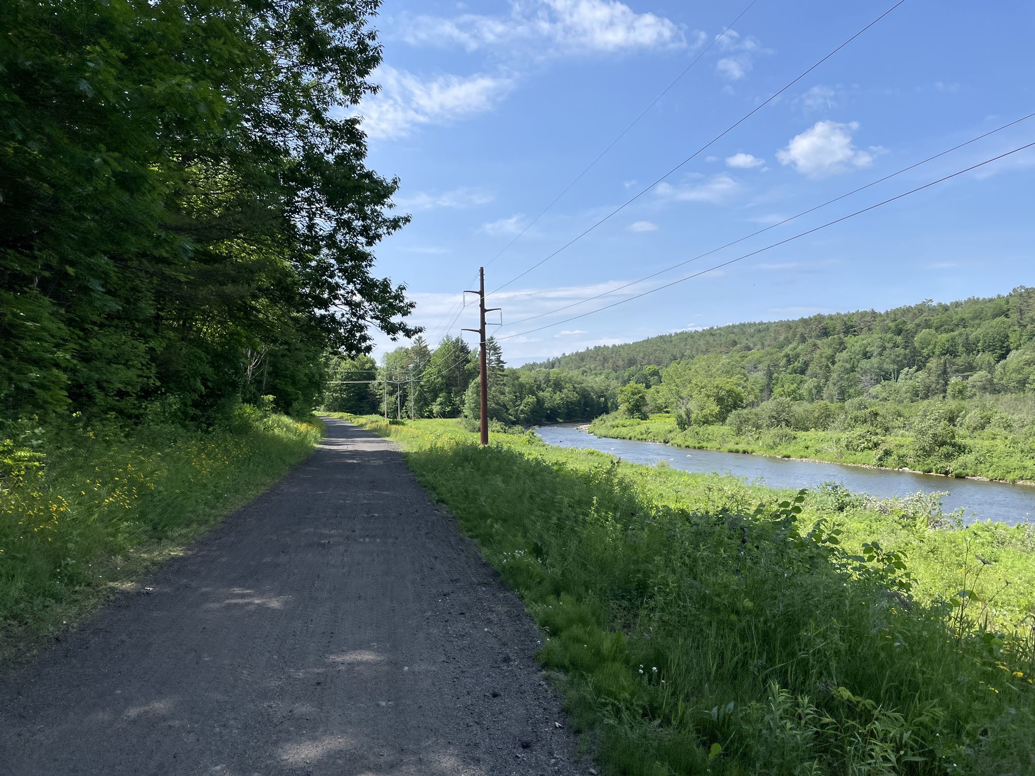 Gravel path alongside a river with forested hills under a sunny sky. - Cross New Hampshire Adventure Trail