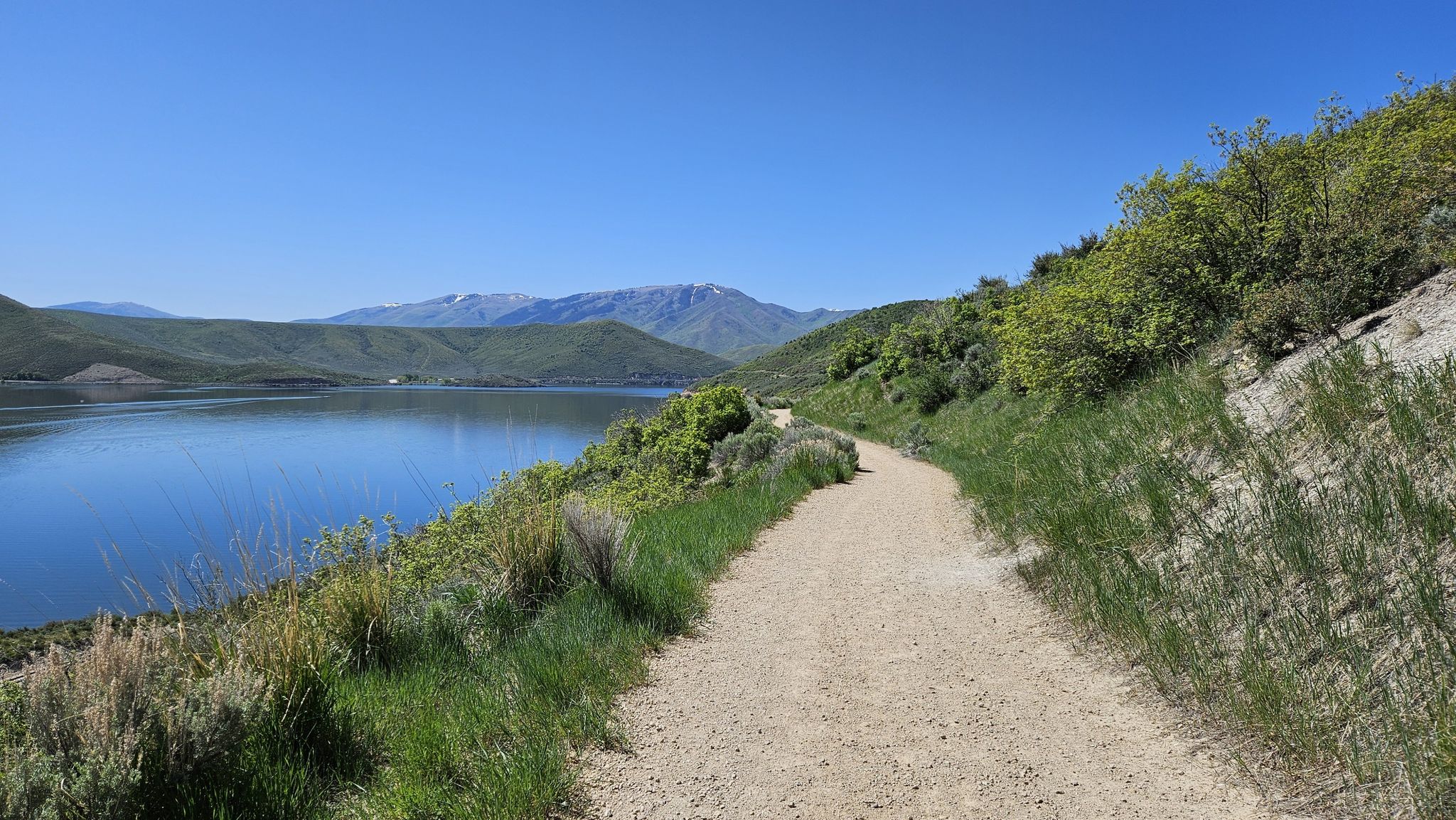 A dirt path follows a large lake with green hills and distant mountains. - Deer Creek Trail