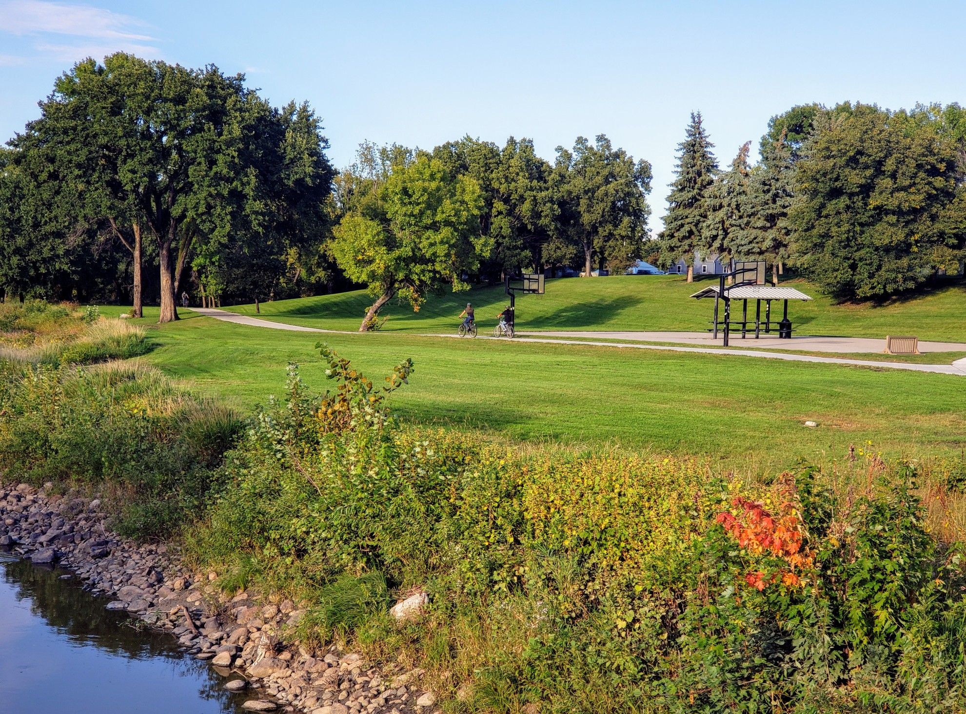 Two people cycling on a path in a sunny green park with a gazebo. - Bergquist Pioneer Cabin – Red River Trail loop from Concordia College