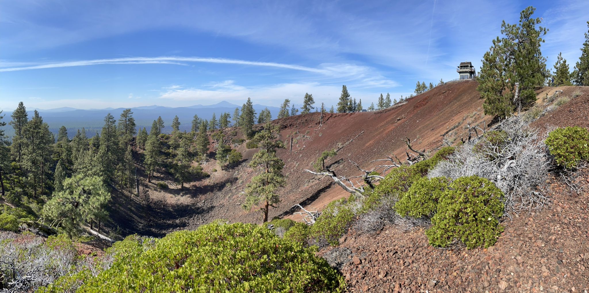 Panoramic view of a red volcanic slope with pine trees and a distant lookout tower. - Lava Butte Trail Loop