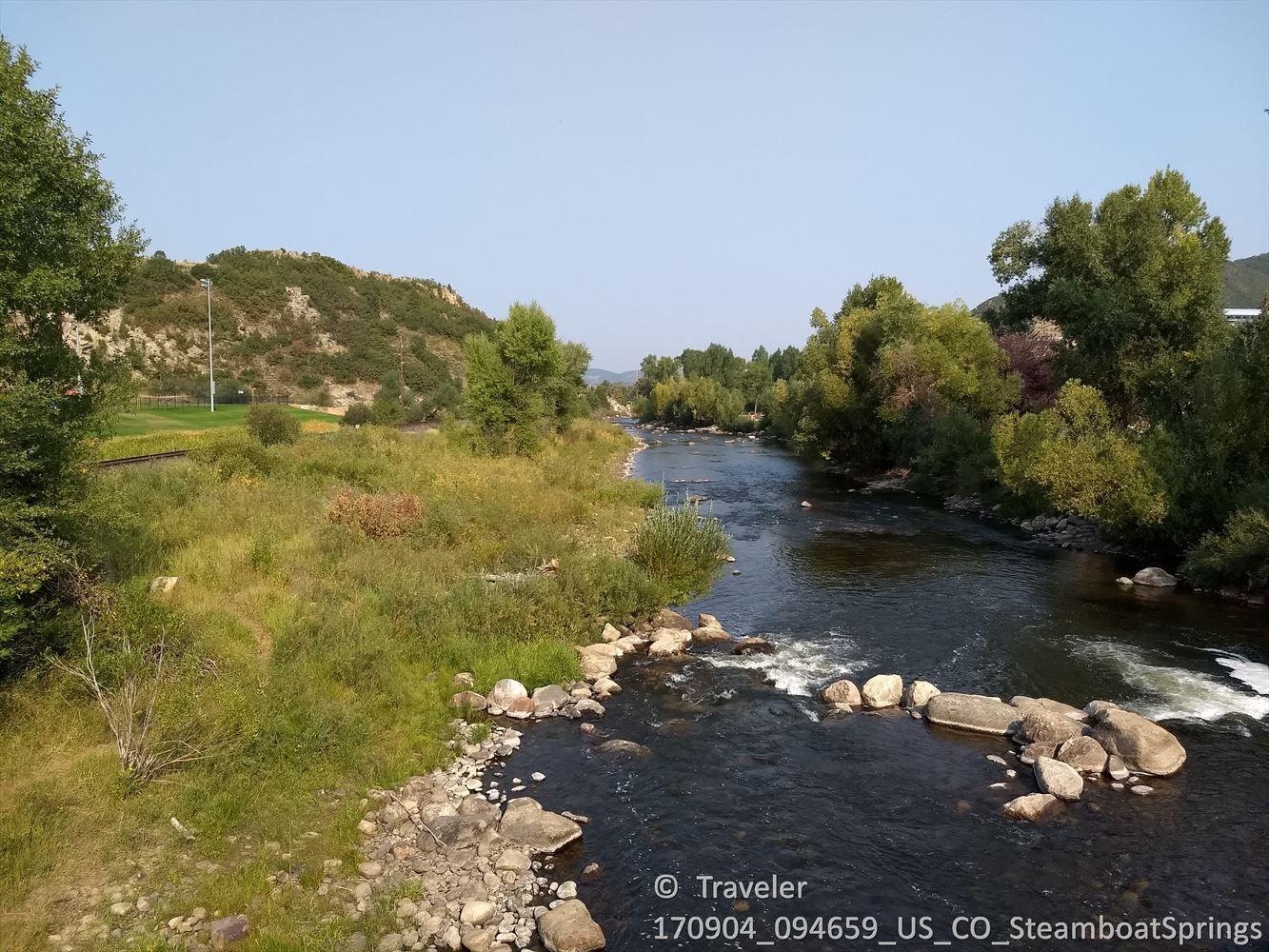 River flows past rocky banks and green hills under a clear blue sky. - Steamboat Springs, Colorado – Orange Peel Bike Shop loop from Steamboat Springs