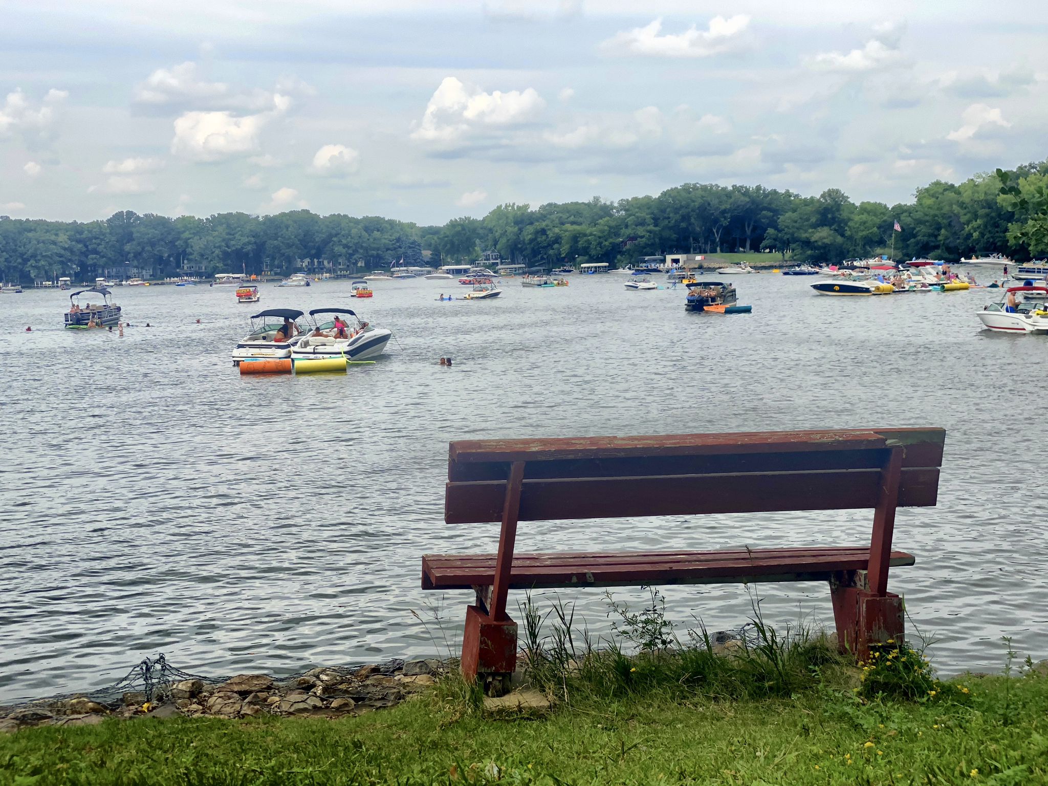A wooden bench on a grassy bank overlooks a lake with numerous boats. - Maywood Beach – Wheelers Beach loop from Spirit Lake