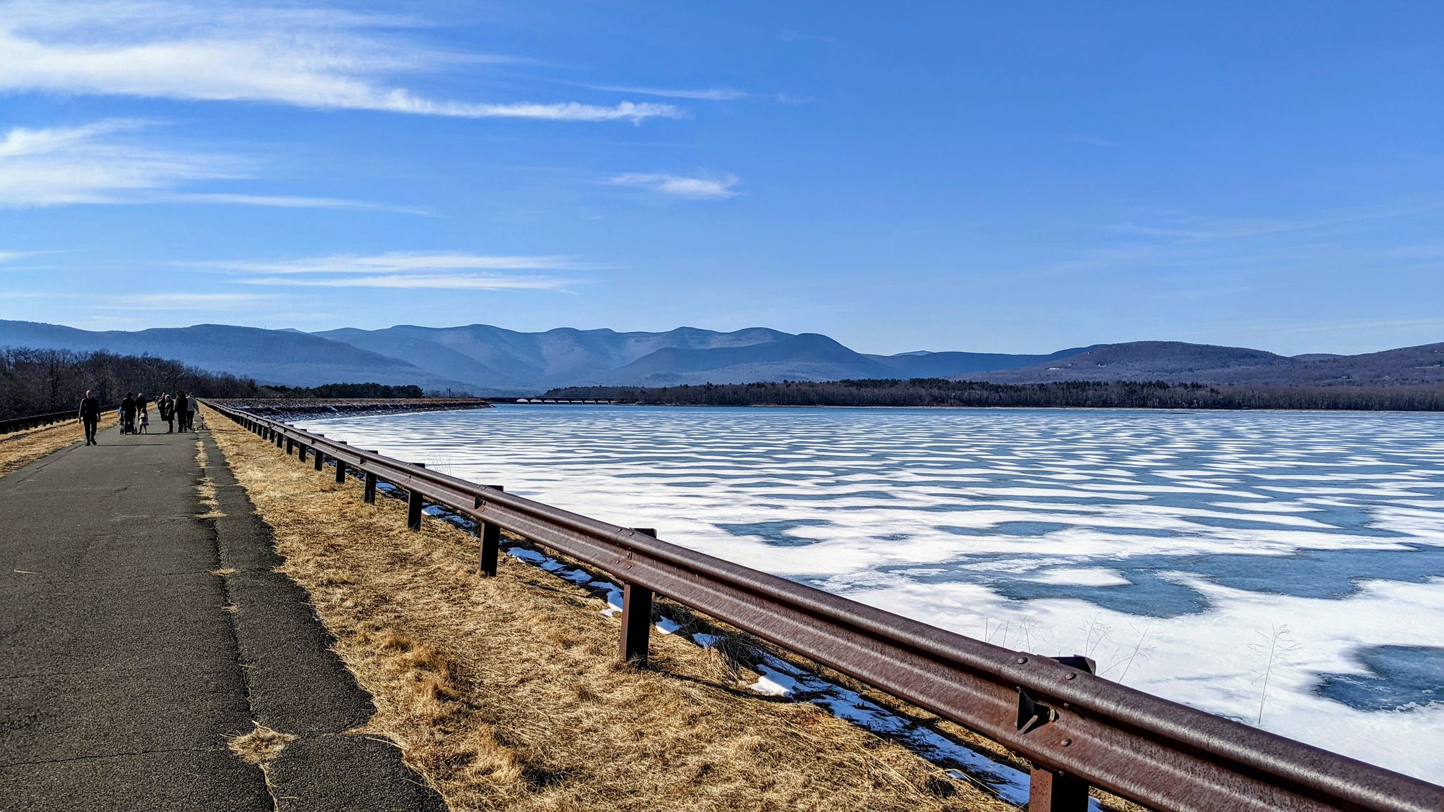 People walk on a paved road beside a partially frozen lake, distant mountains. - Ashokan Reservoir Promenade via BWS Road