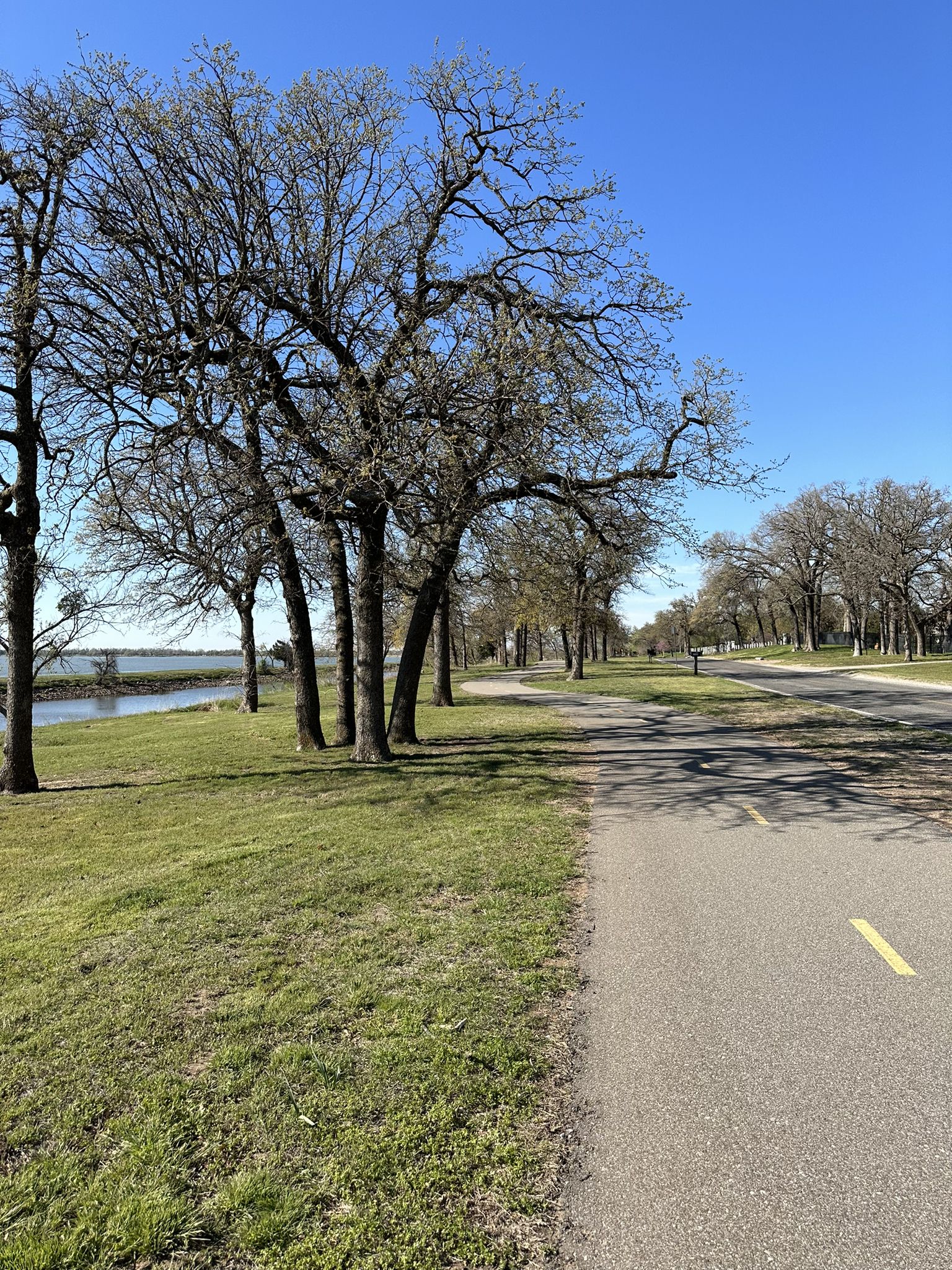 Paved path lined with bare trees next to water under blue sky. - Lake Overholser Dam – Lake Overholser Bridge loop from Amazon OKC5