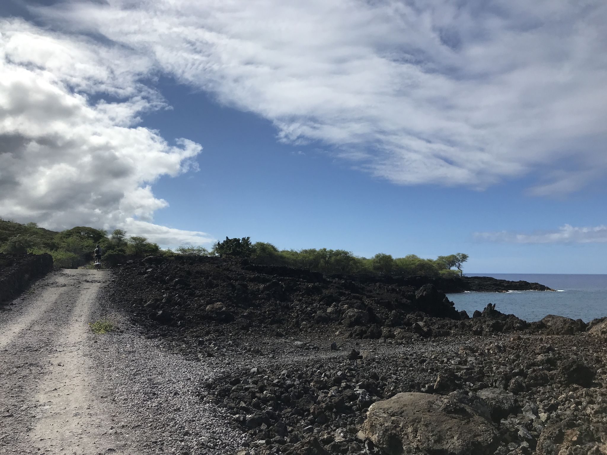 Gravel road curves left, dark volcanic rock, ocean, and cloudy sky. - End of the World Trail