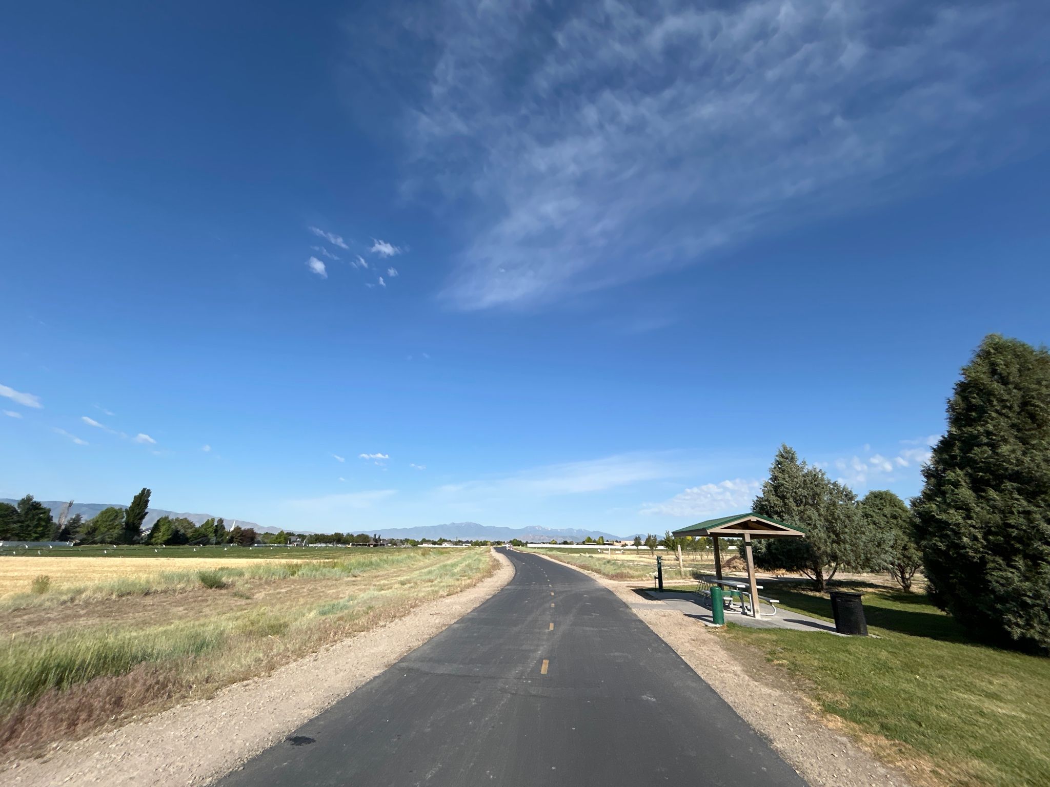 Paved path through green fields, leading to a picnic shelter under blue sky. - Murdock Canal Trail