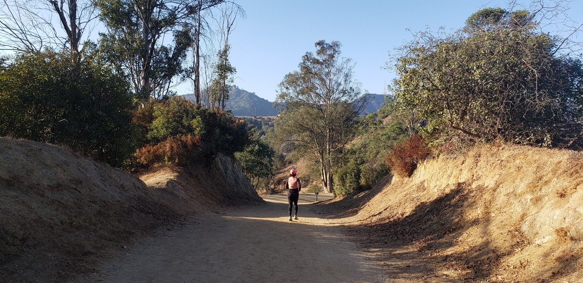 Person walks on a sunny dirt path through dry hills and trees. - Condor, Skyline and Rattlesnake Trail