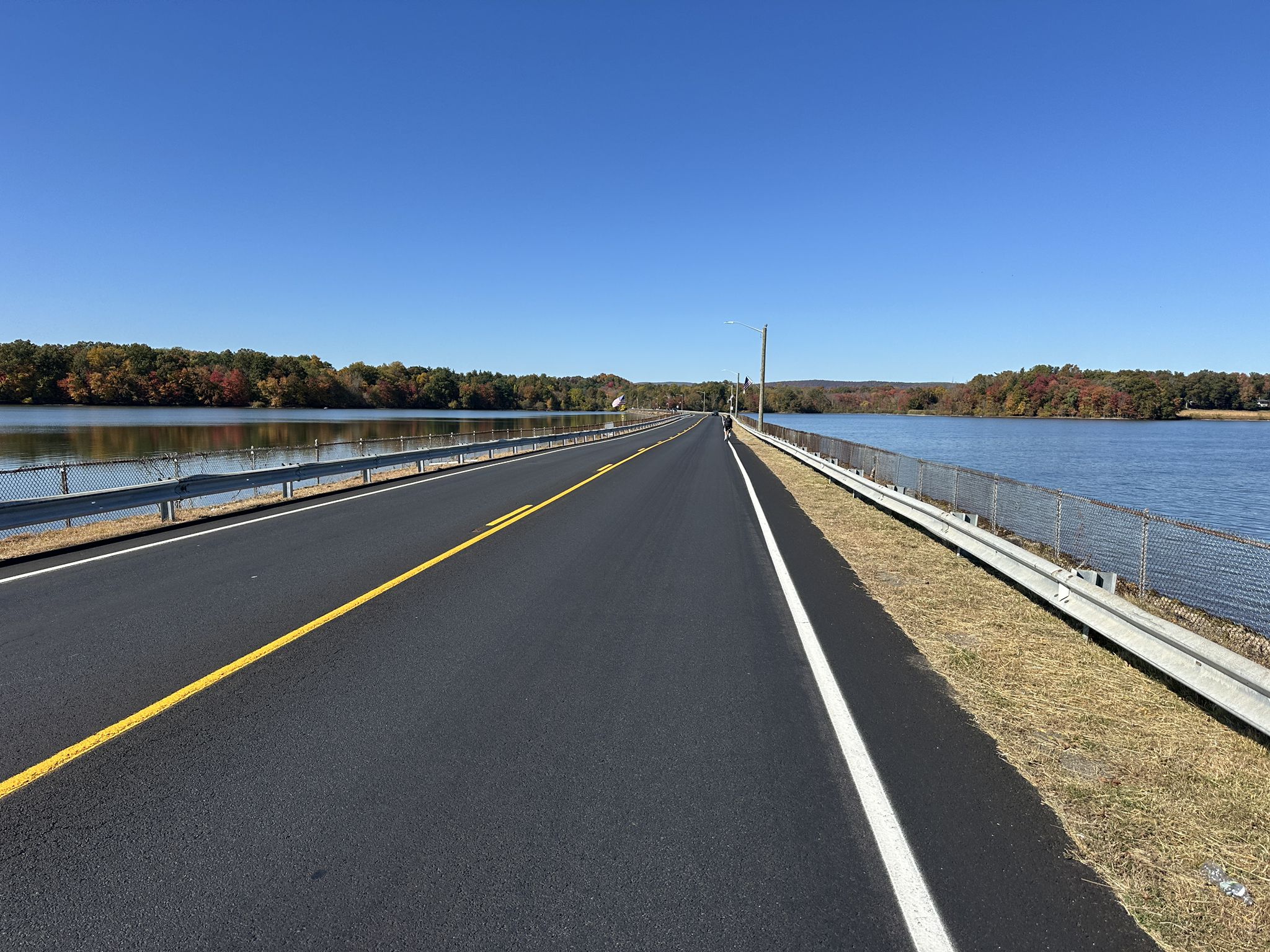 Long road over water, flanked by fences and trees, under a clear blue sky. - Rockland Lake State Park Loop