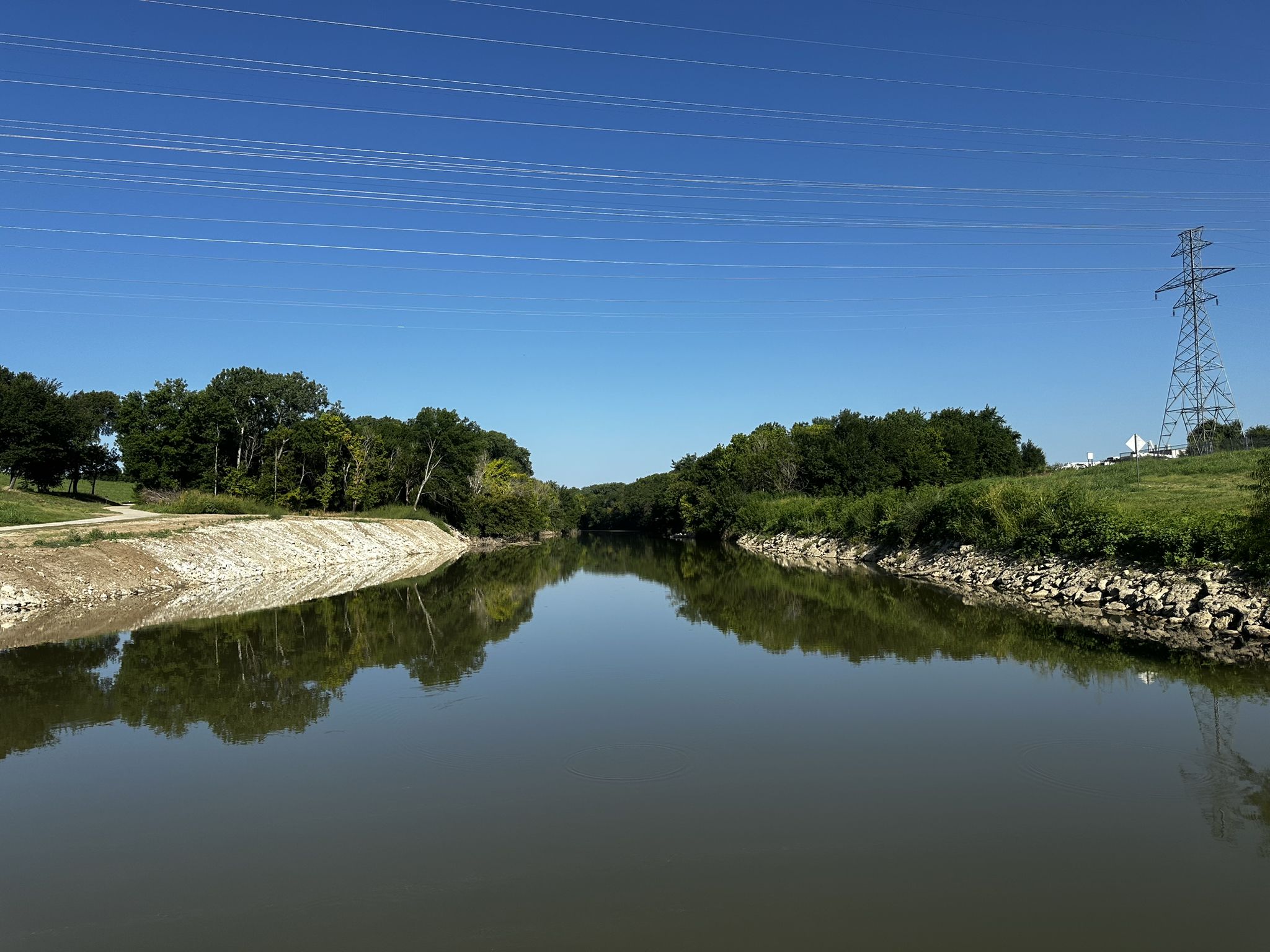 A wide, calm river reflects trees and a clear blue sky. - Harmon Field Park – Trinity River loop from Trinity Lakes Station