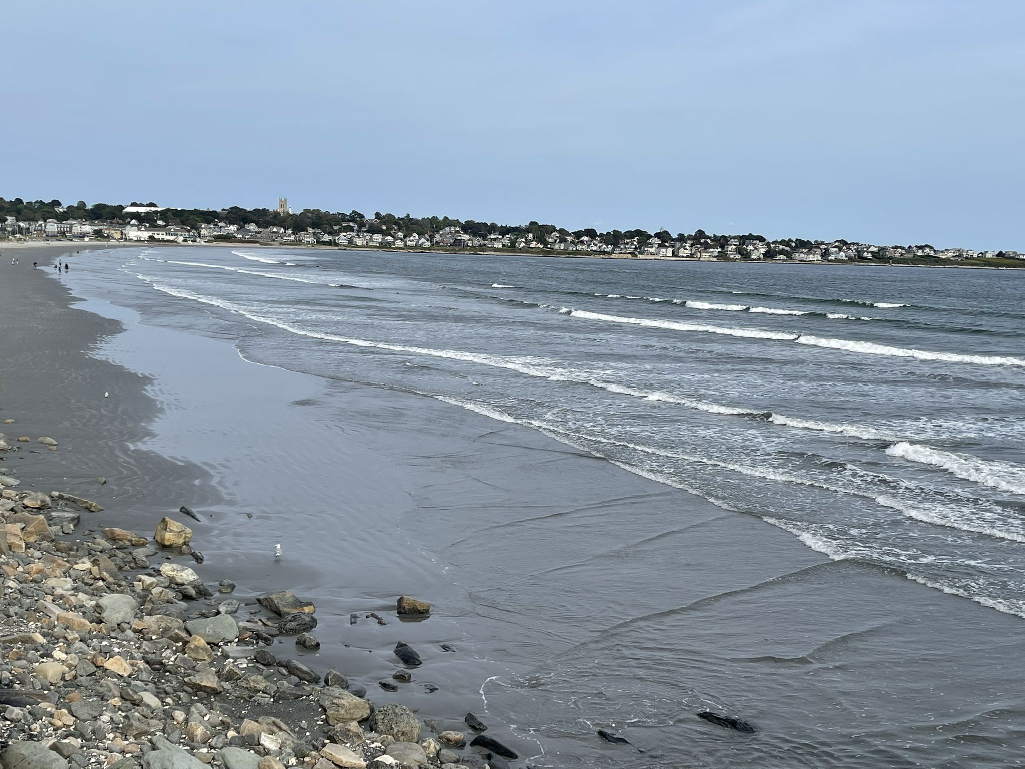 Overcast day at a wide sandy beach with gentle waves and a distant town. - Perroti Park and Marina – Museum of Newport History loop from Newport