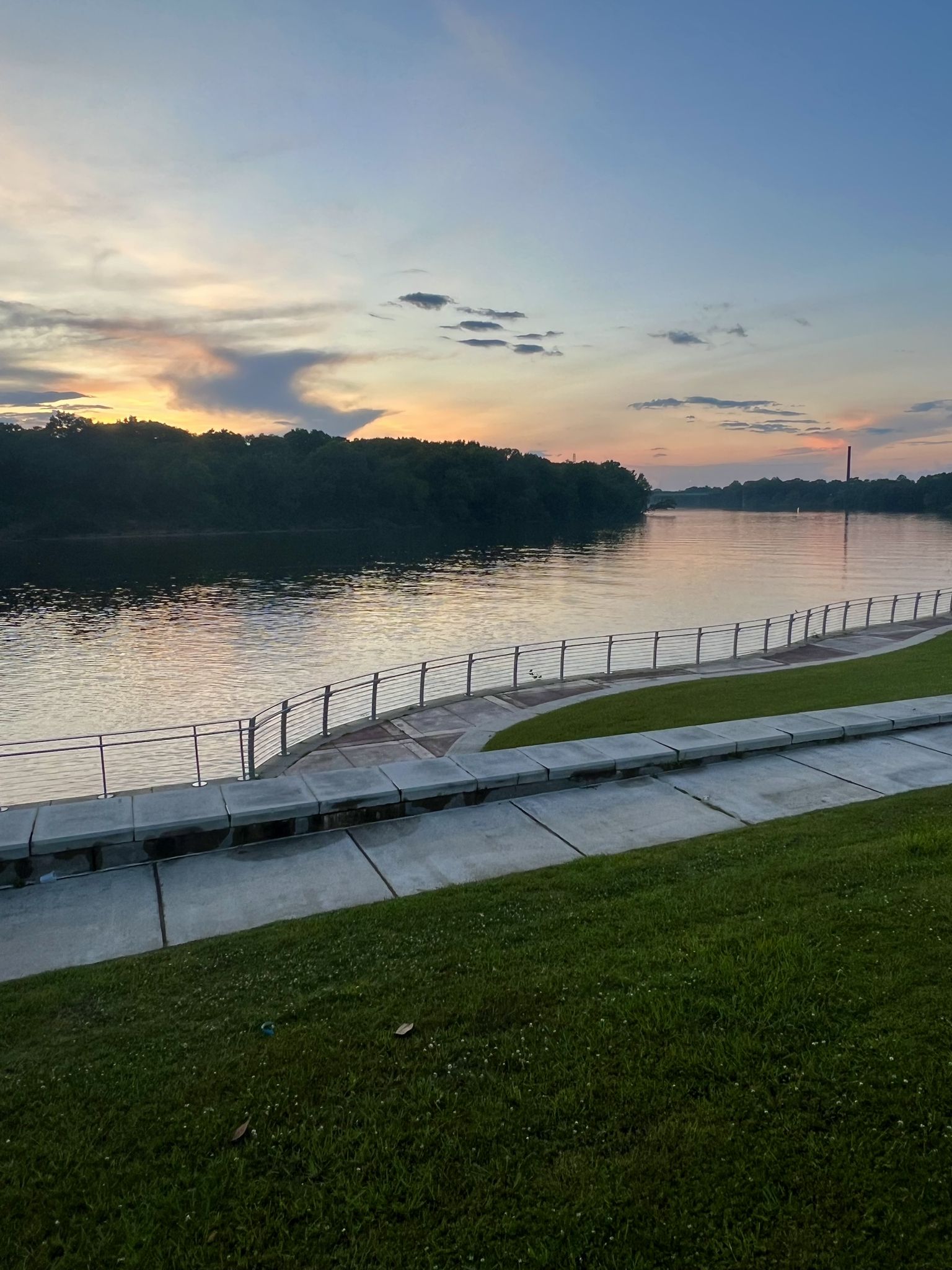 Sunset over a wide river with a waterfront path and green grassy bank. - Riverfront Greenway Trail