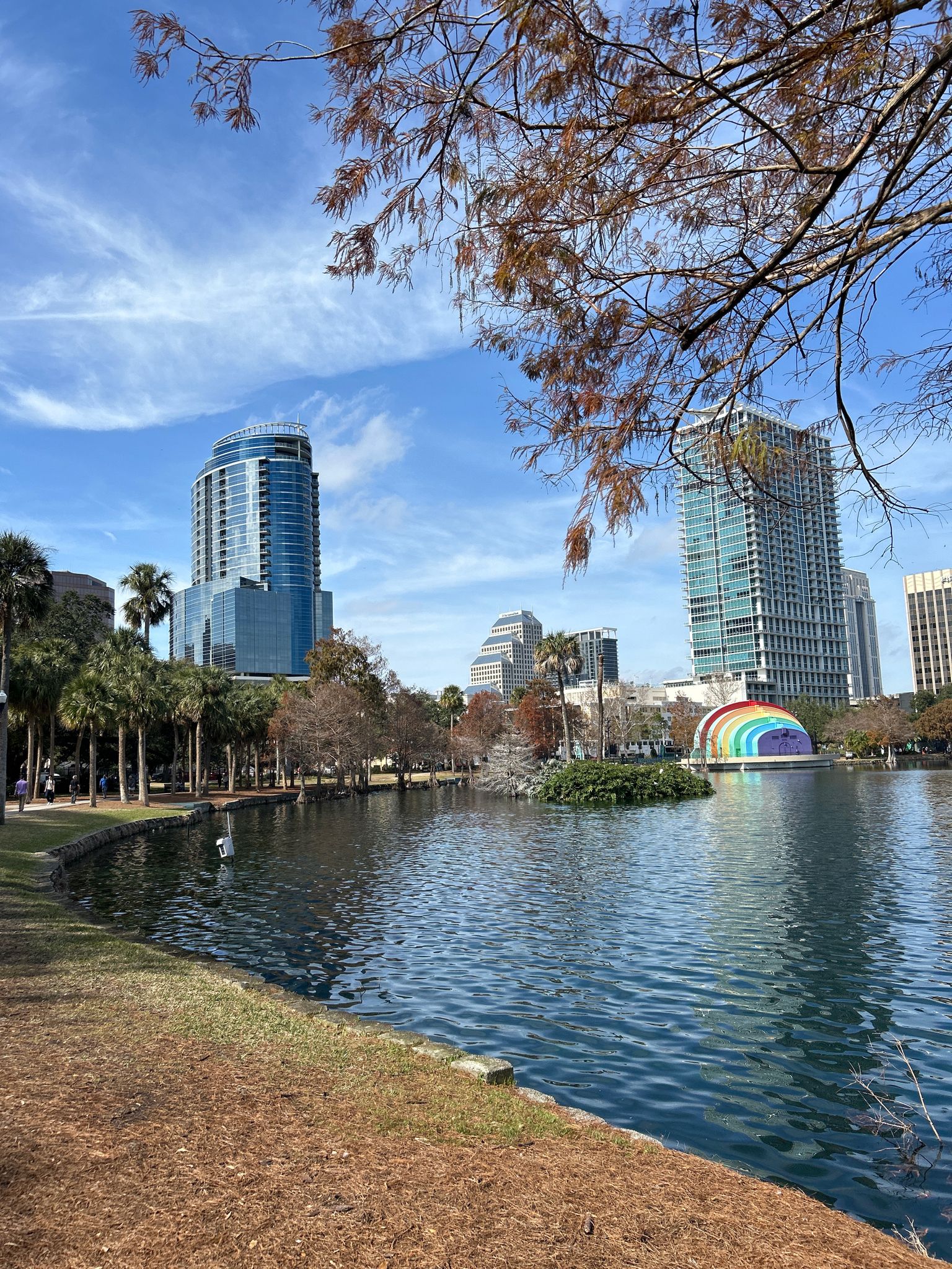 Lake with a colorful rainbow structure, city buildings, and trees under blue sky. - Lake Eola Park Loop – Lake Eola Fountain loop from Orlando