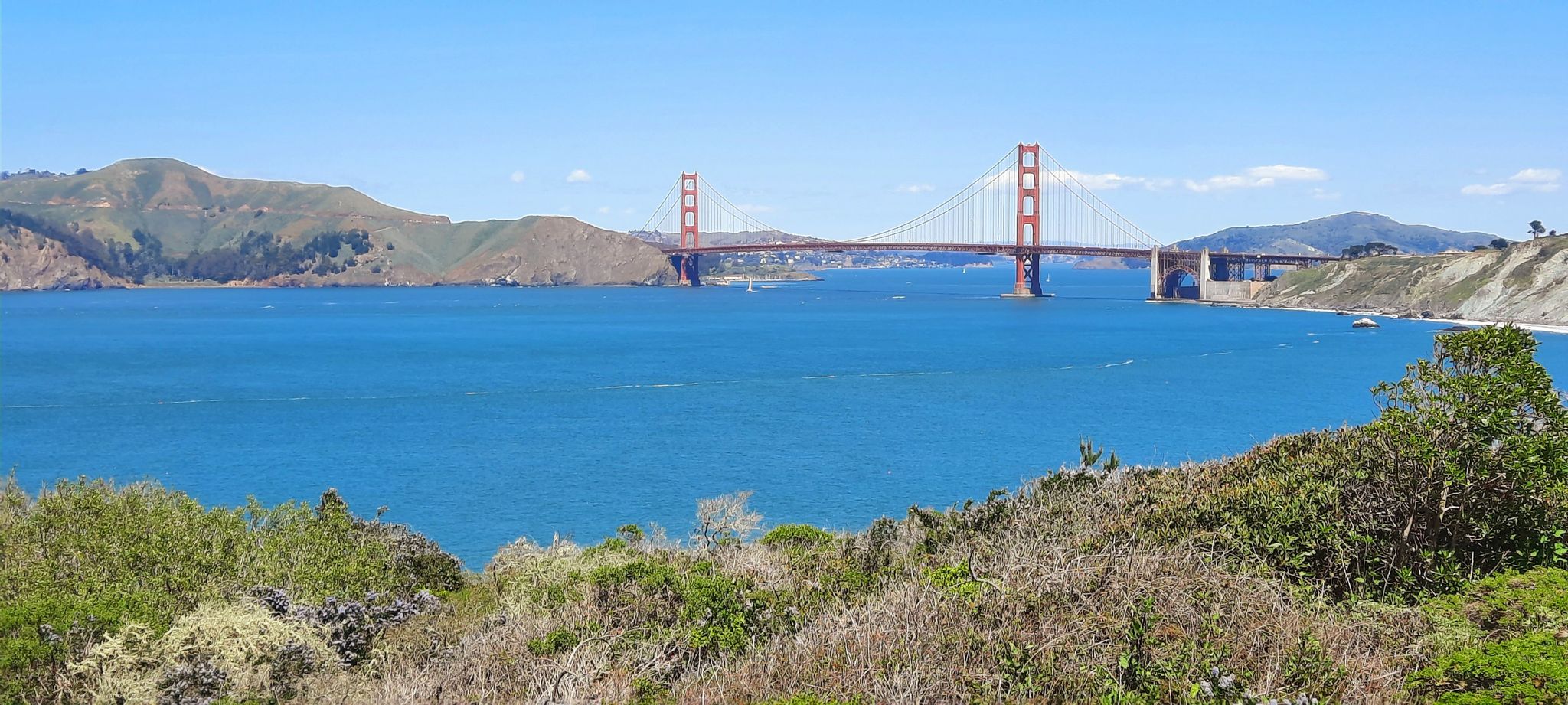 Red suspension bridge over wide blue bay, green hills, sunny. - Coastal Trail