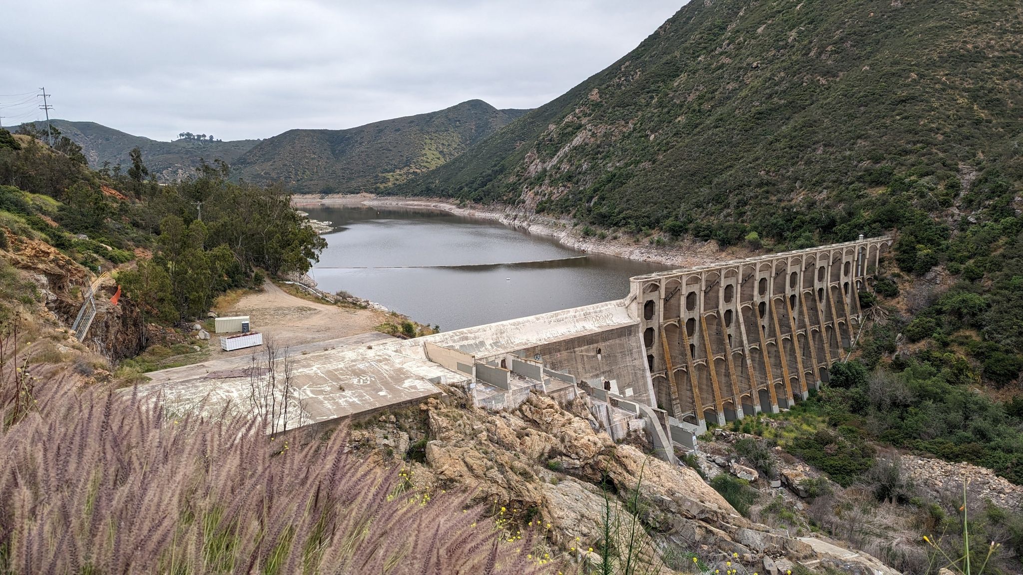 Overcast view of a large concrete dam holding back a reservoir, surrounded by hills. - Hodges Dam – Lake Hodges Trail loop from San Elijo Hills