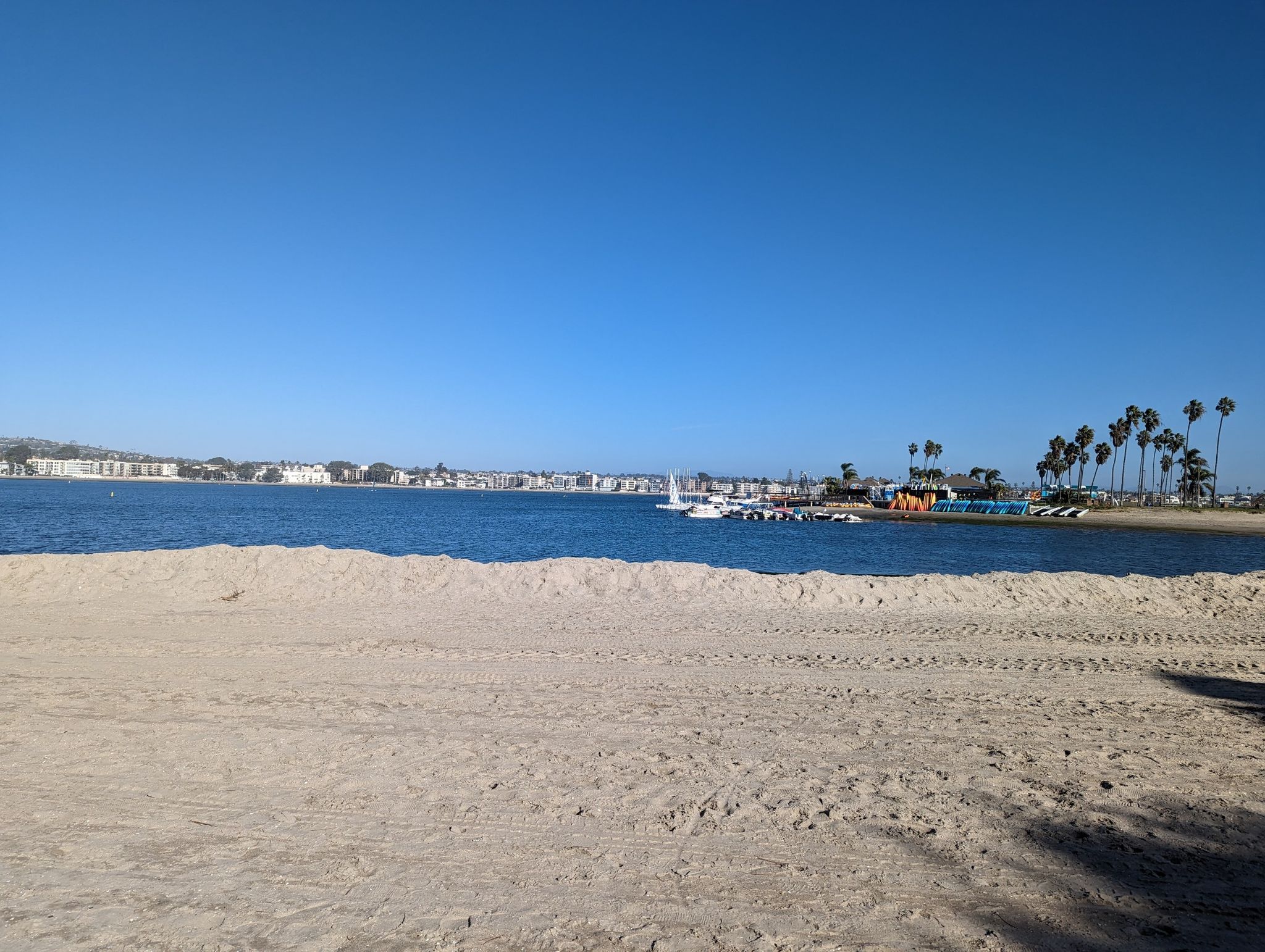 A wide, empty sandy beach stretches towards calm water with distant buildings under a clear blue sky. - SDSU Beach – Mission Bay loop from Pacific Beach