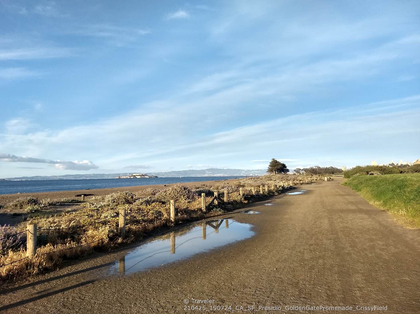 Dirt path with puddles next to a fenced beach and calm ocean under blue sky. - Golden Gate Promenade – Marina Yacht harbor ⛵ loop from Presidio of San Francisco