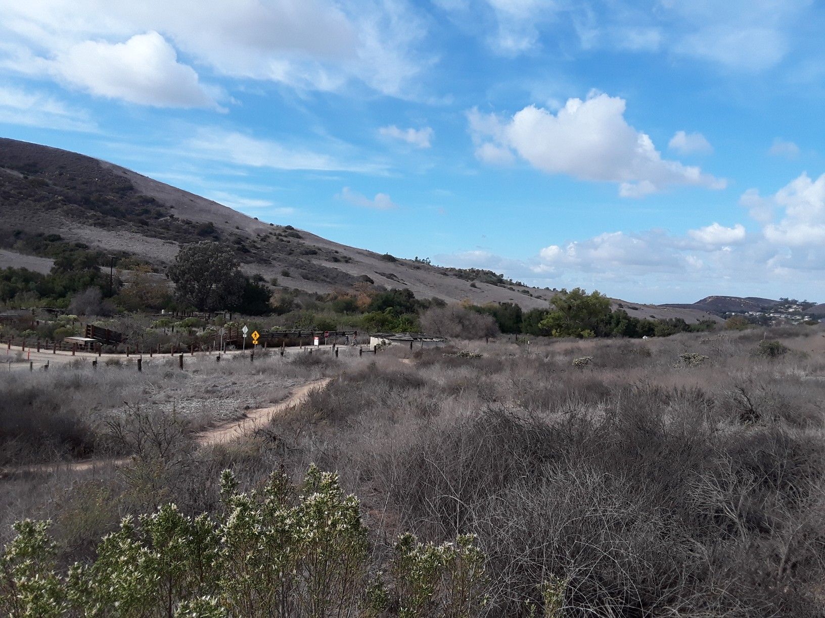 Dirt path next to a road, dry brush, and hills under a partly cloudy sky. - Walnut Trail – Shady Canyon Trail loop from Tustin