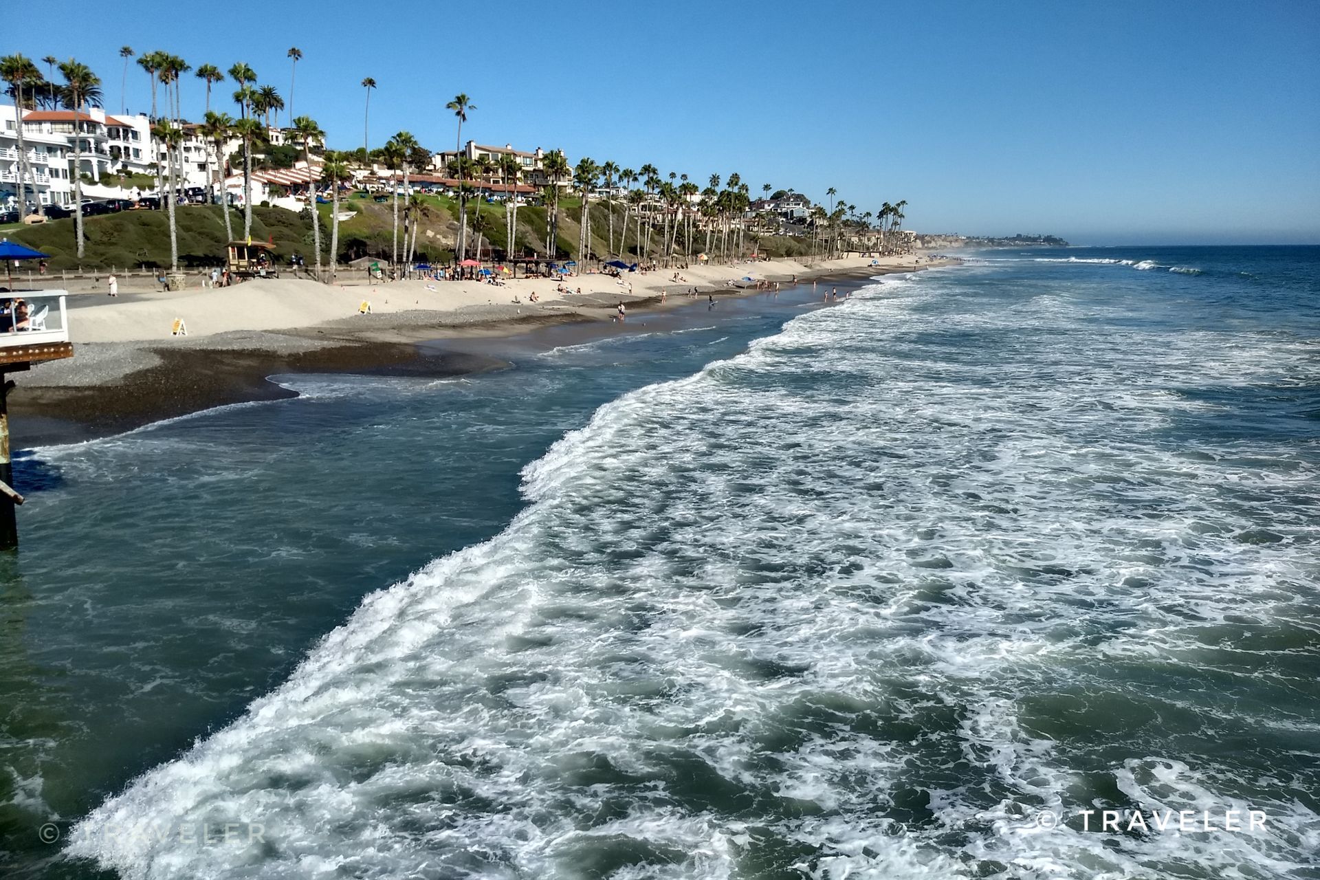 Sunny beach with crashing waves, palm trees, and coastal buildings. - San Clemente Beach Trail