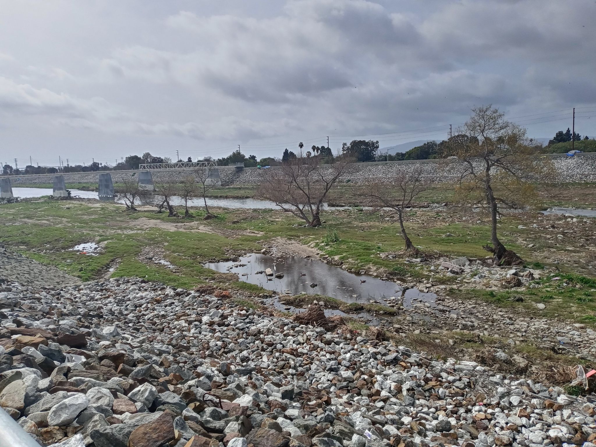 Rocky riverbed with small water pools, sparse trees, and distant bridge. - Redondo Beach – RMS Queen Mary loop from Redondo Beach