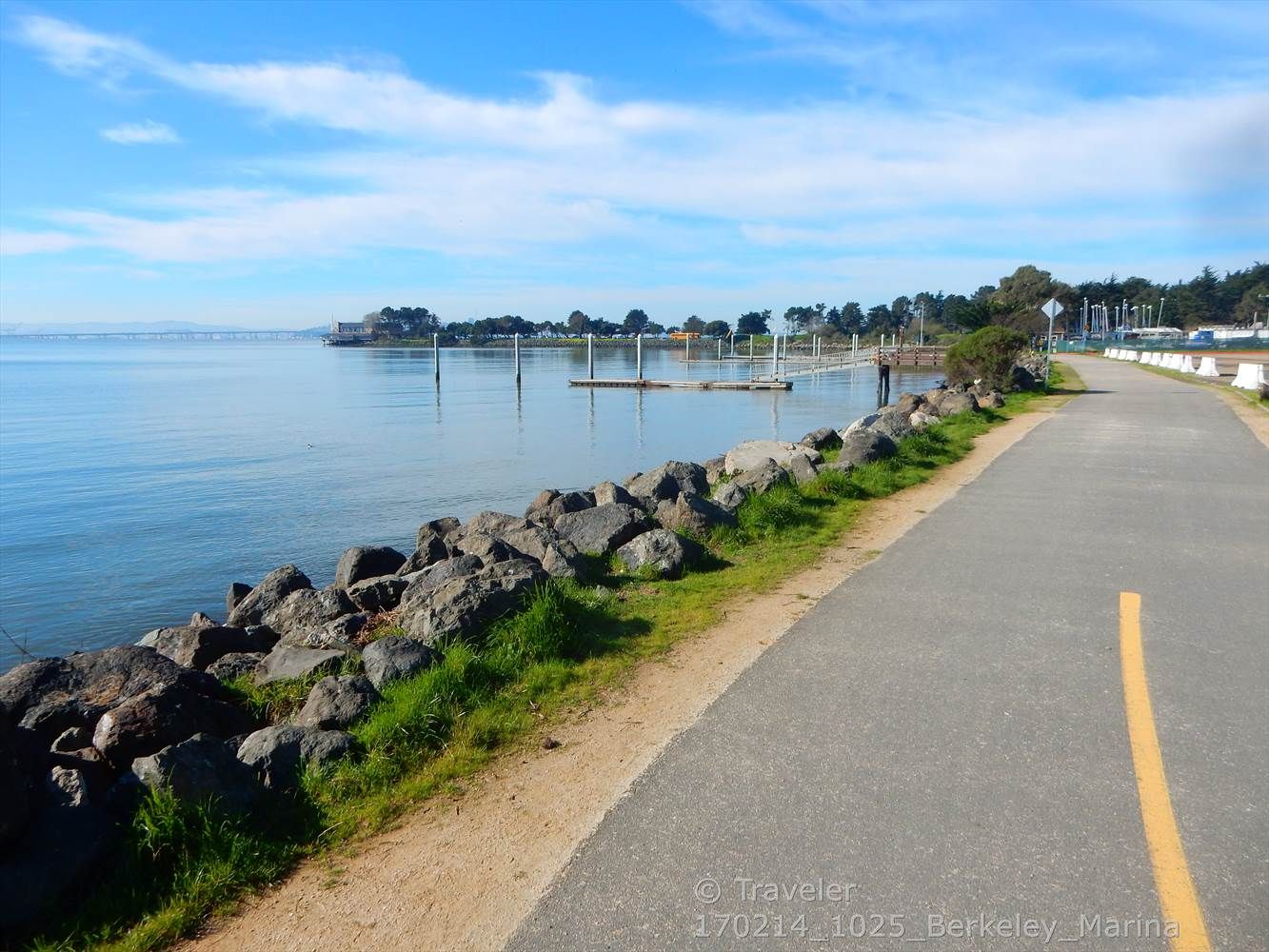 Paved path beside rocky shore, calm water with docks, under a bright blue sky. - West Street Bikeway – Ohlone Greenway loop from El Cerrito del Norte