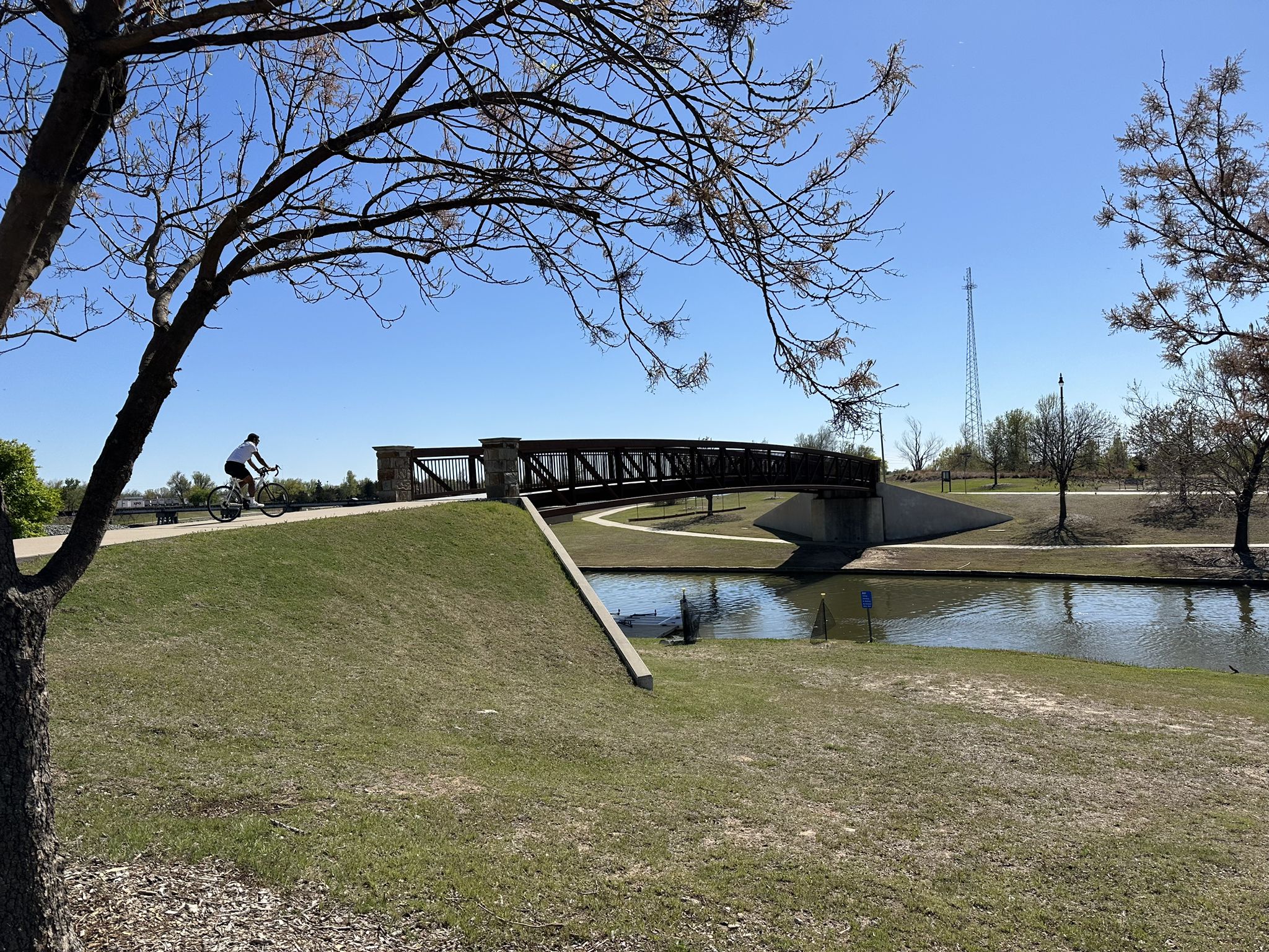 Cyclist on a paved path approaching a pedestrian bridge over water. - Oklahoma River Trail: Bricktown Loop
