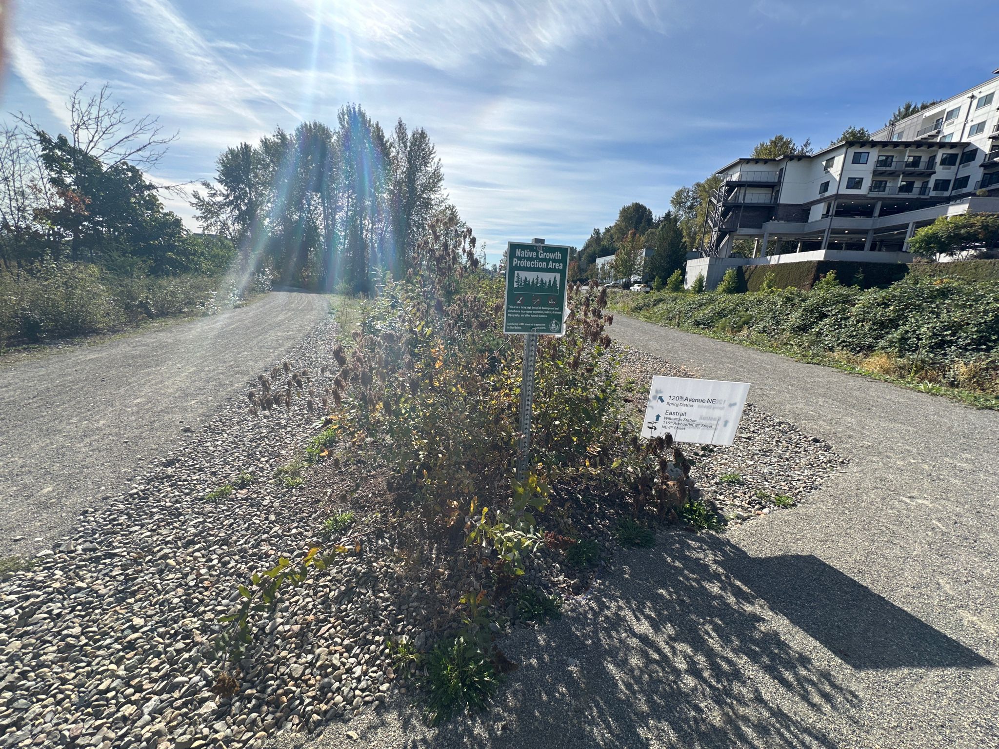 Sunny gravel path with signs, bushes, and distant buildings on a hill. - Eastrail to Ashwood Park