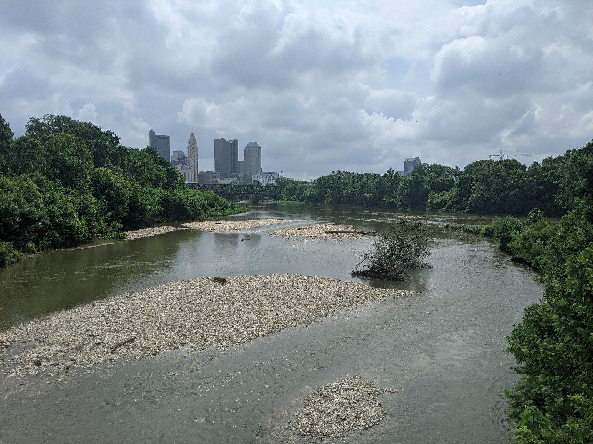 River with gravel bars, green trees, and a distant city skyline under cloudy sky. - North Bank Park Pavilion – Olentangy River loop from Columbus